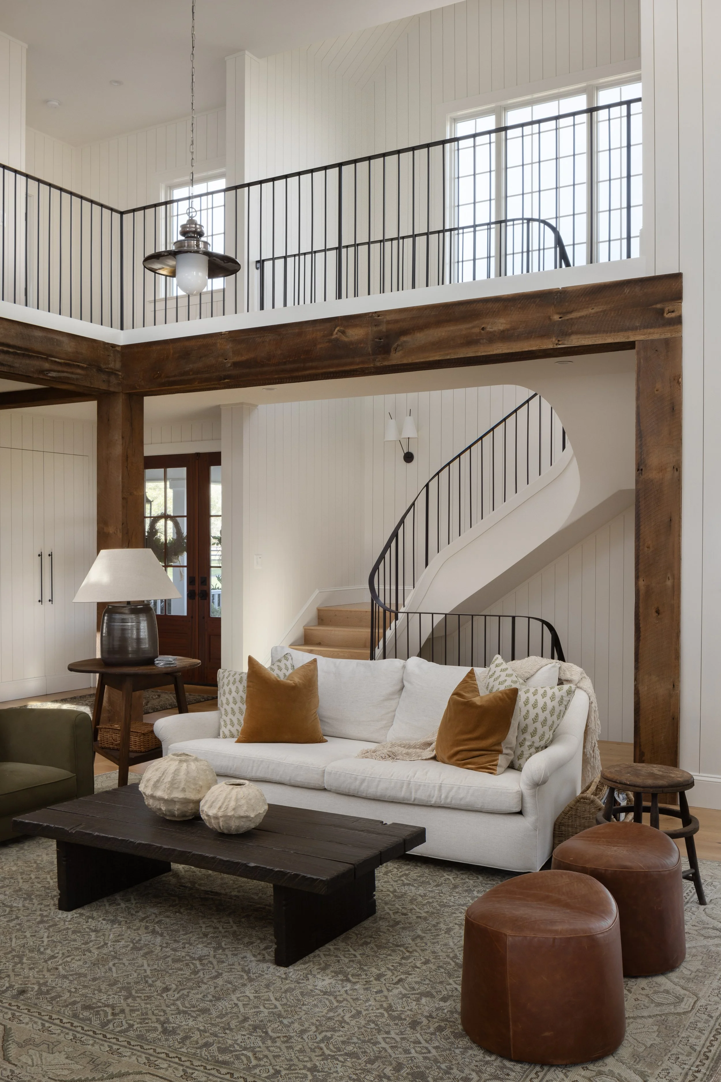Living room with a white sofa, brown pillows, a dark wooden coffee table, a patterned rug, leather poufs, and a side table with a lamp. A staircase with black railings and a dark wood beam is visible in a high-ceiling space with white walls and large