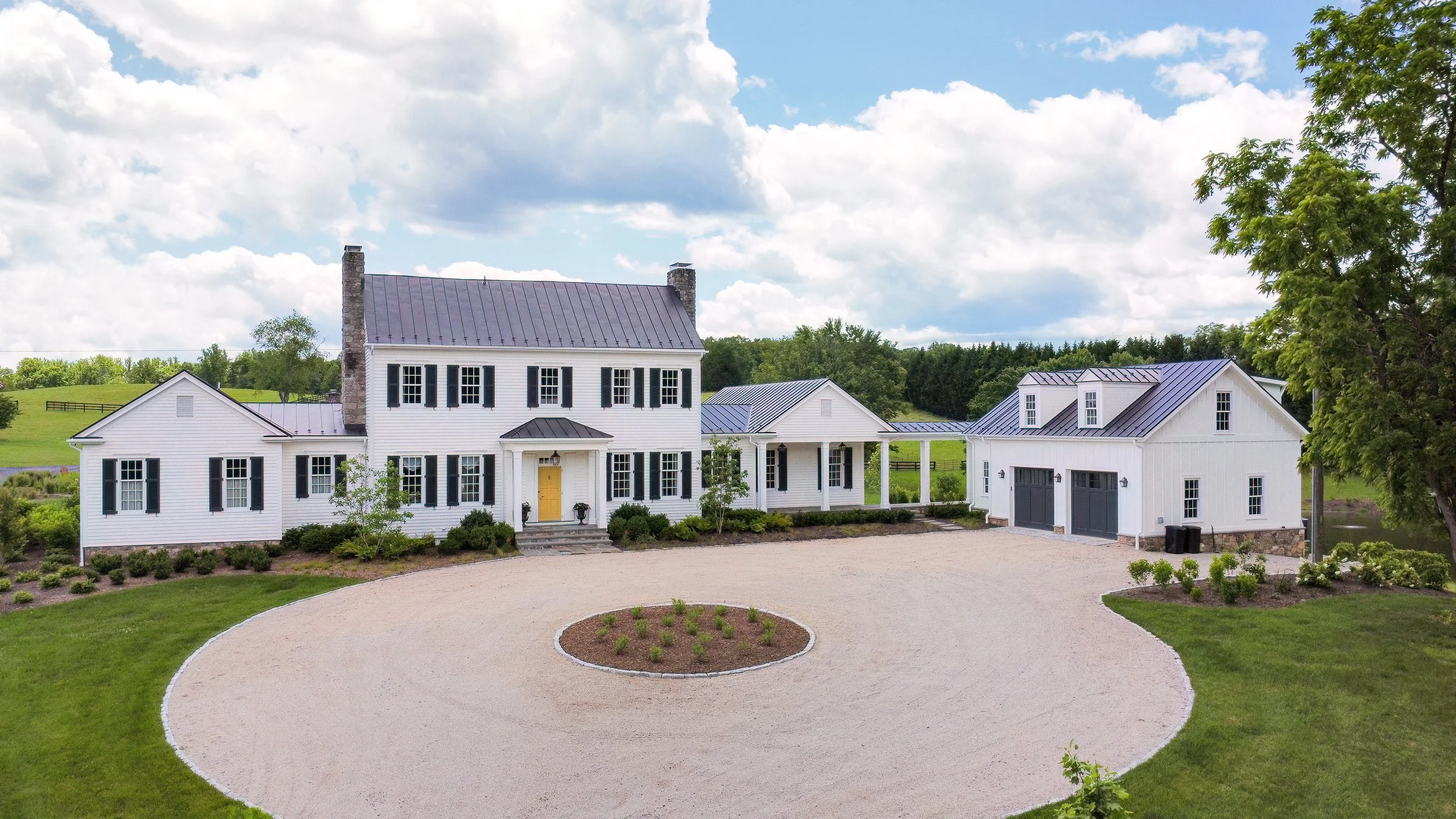 A large white farmhouse with black shutters and a yellow front door, accompanied by a separate garage building, all surrounded by a manicured lawn and a scenic rural landscape with greenery and cloudy sky.