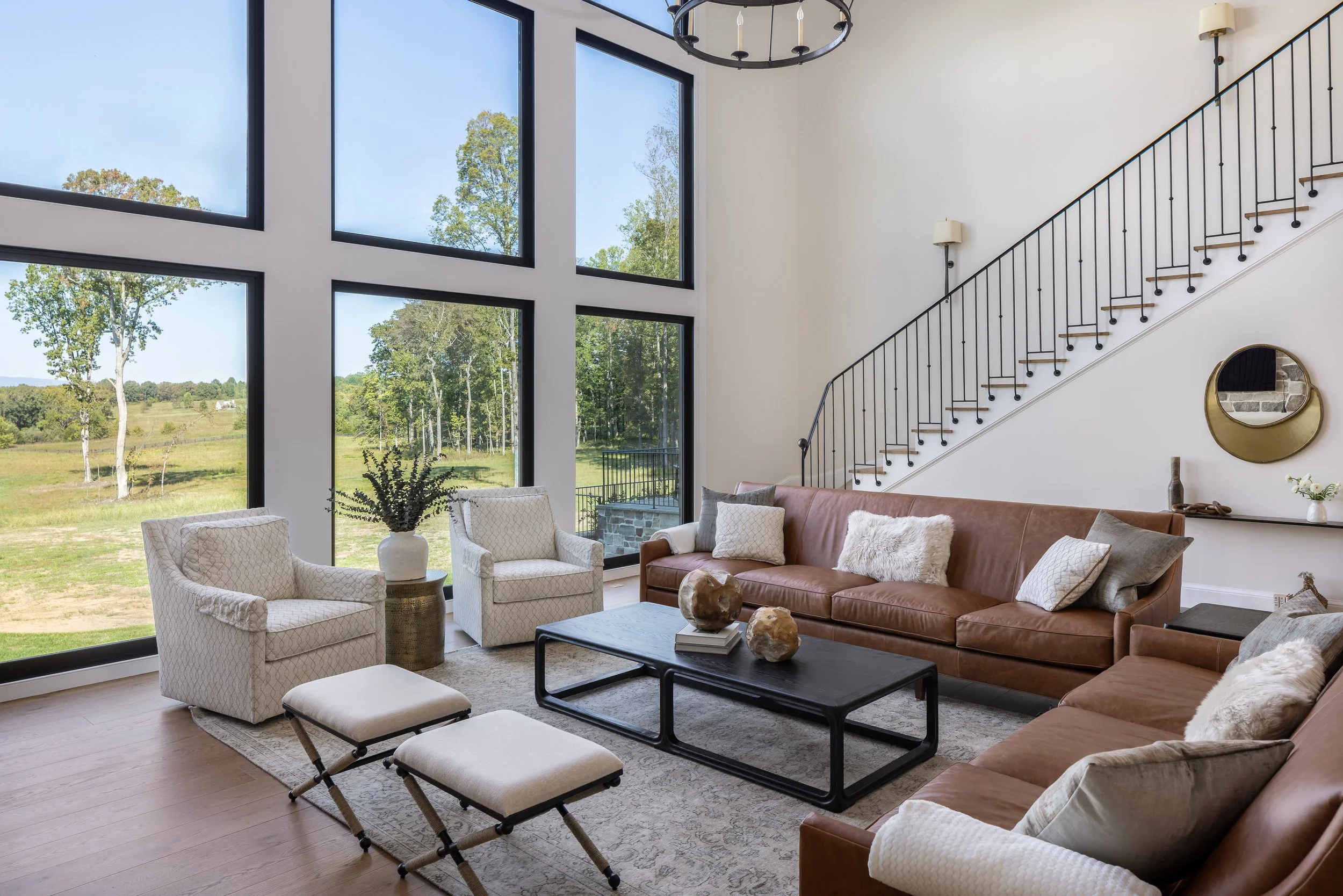 Living room with large windows showing green landscape, featuring tan leather sofa, white armchairs, and a black coffee table decorated with decorative stones, pillows, and a plant.