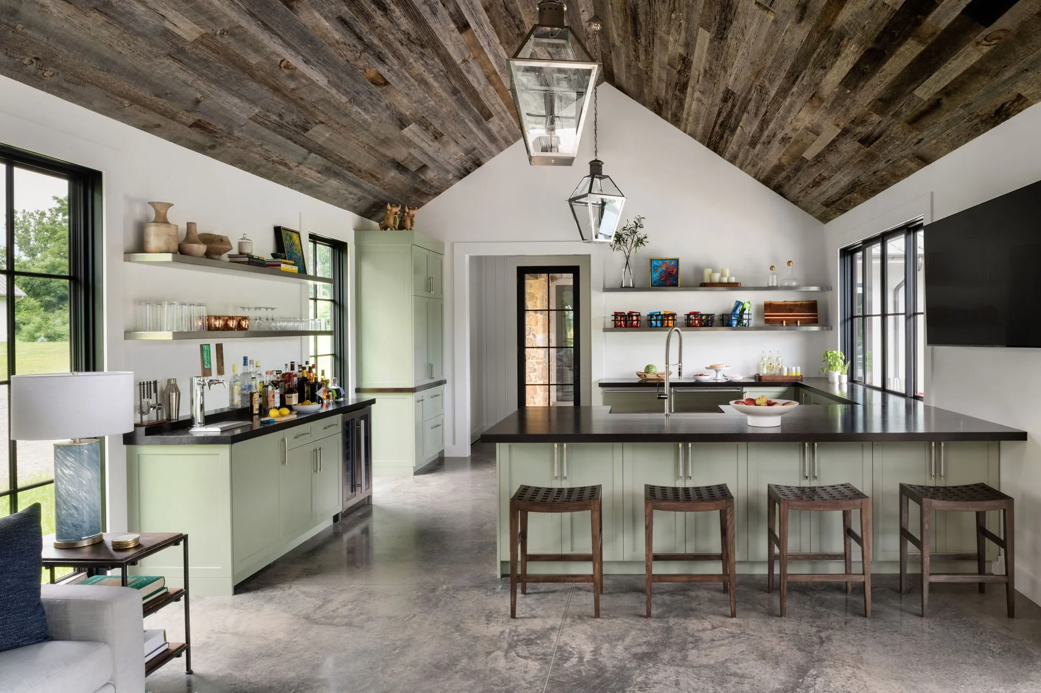 Modern kitchen with high vaulted wooden ceiling, mint green cabinets, a black countertop, a kitchen island with stools, open shelves with various kitchen items, and large windows showing greenery outside.