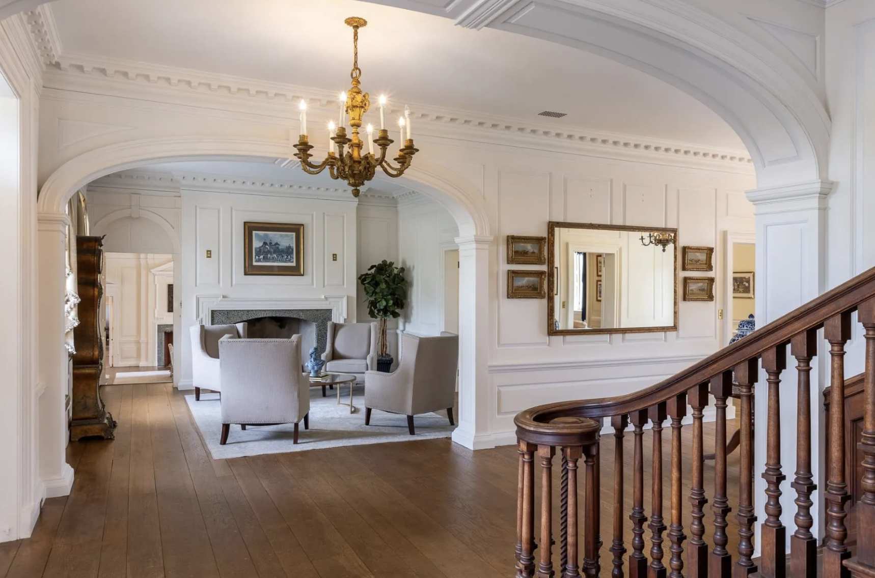 Elegant living room with white walls, a chandelier, hardwood floors, a mirror, paintings, and a seating area with four armchairs around a small glass coffee table.