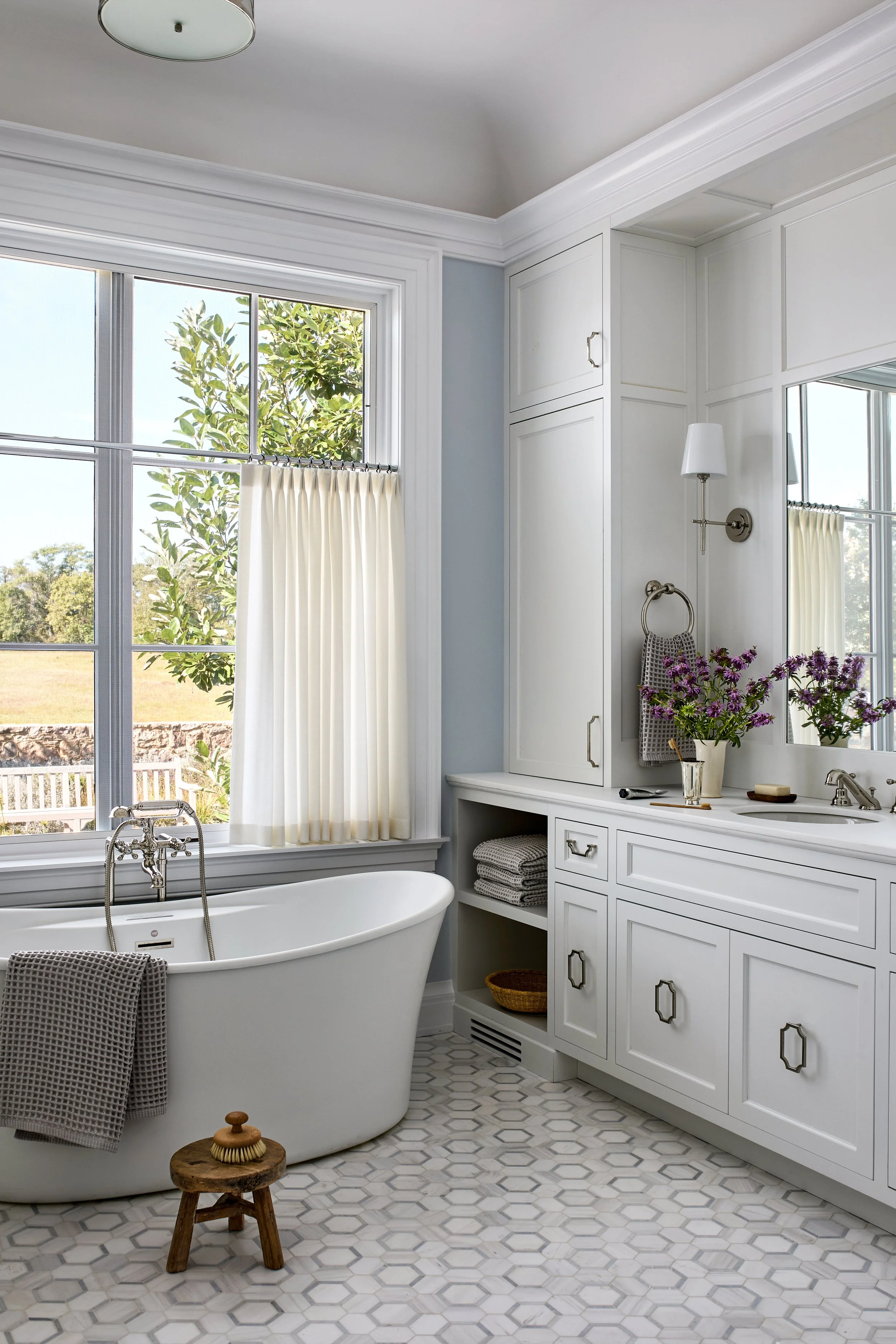 A bathroom with a white freestanding bathtub in front of large windows, white cabinets, a sink with a mirror, and a bouquet of purple flowers on the countertop.