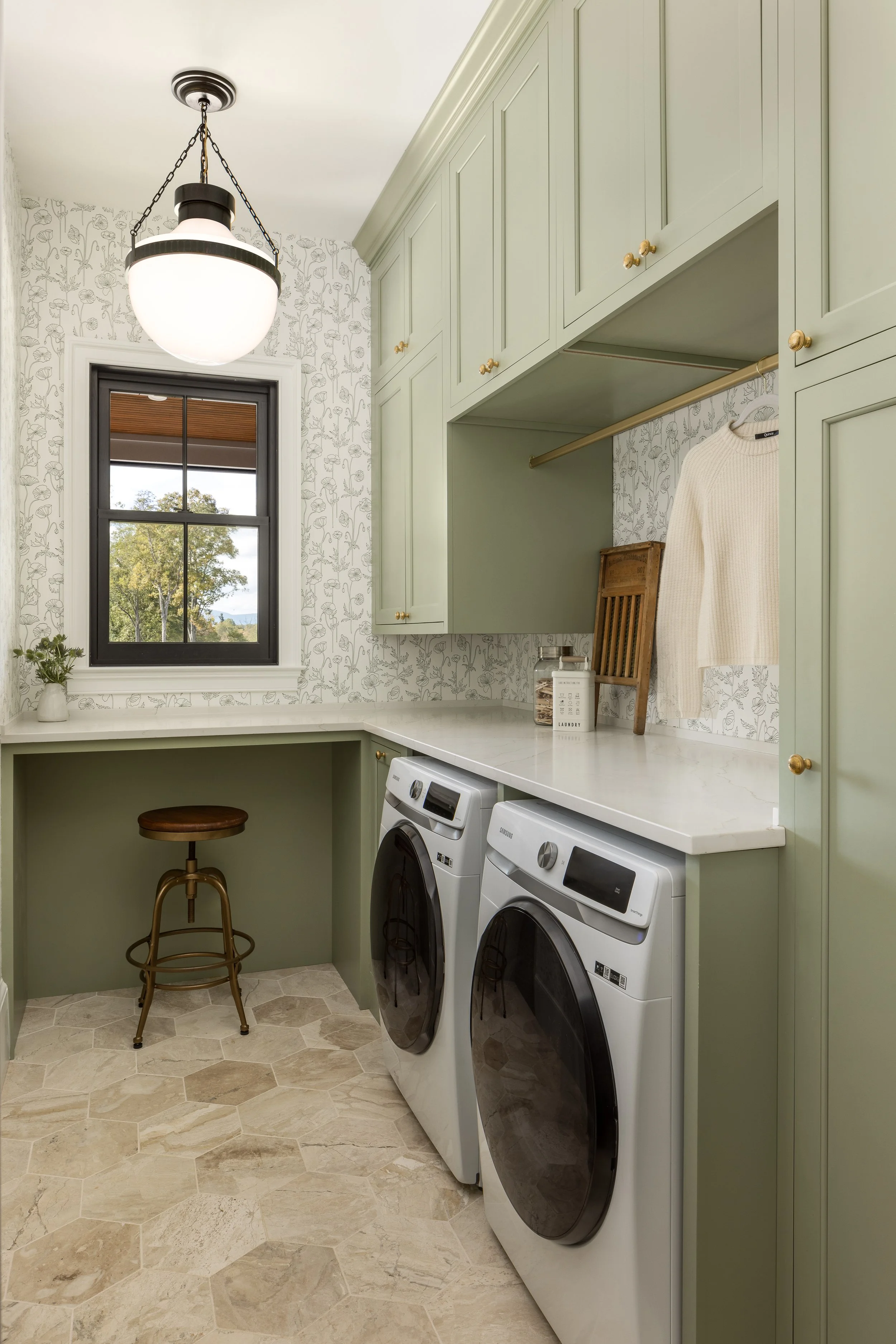 A laundry room with green cabinets, a window showing trees outside, and a white washing machine and dryer. There is a white countertop, a small wooden stool, and a decorative shelf with a jacket hanging. The room has a patterned wallpaper and a moder