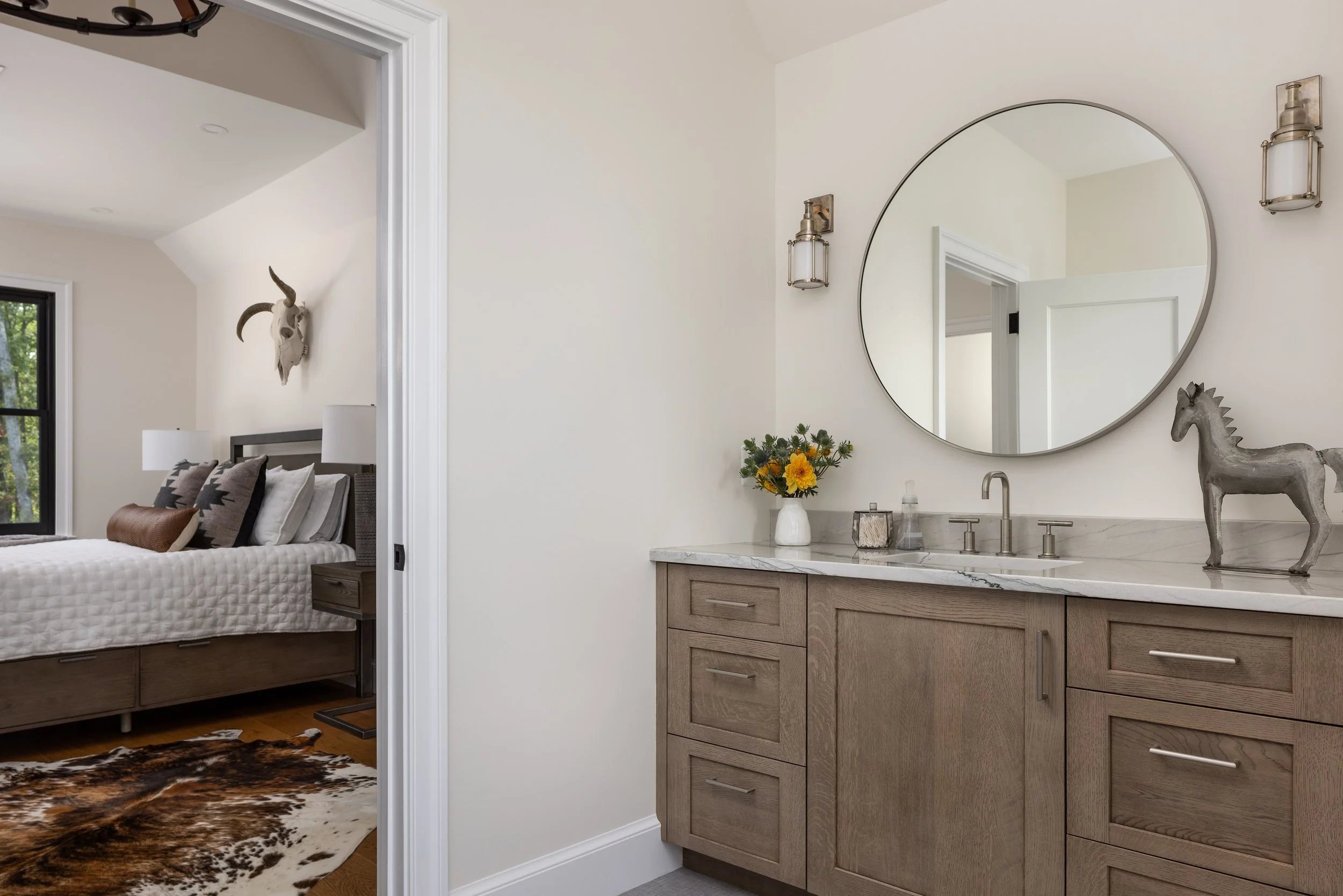 A bathroom vanity with a marble countertop, round mirror, two wall-mounted lights, a decorative horse sculpture, and a vase of yellow flowers. Bedroom visible through doorway with a bed, pillows, and a bull skull on the wall.