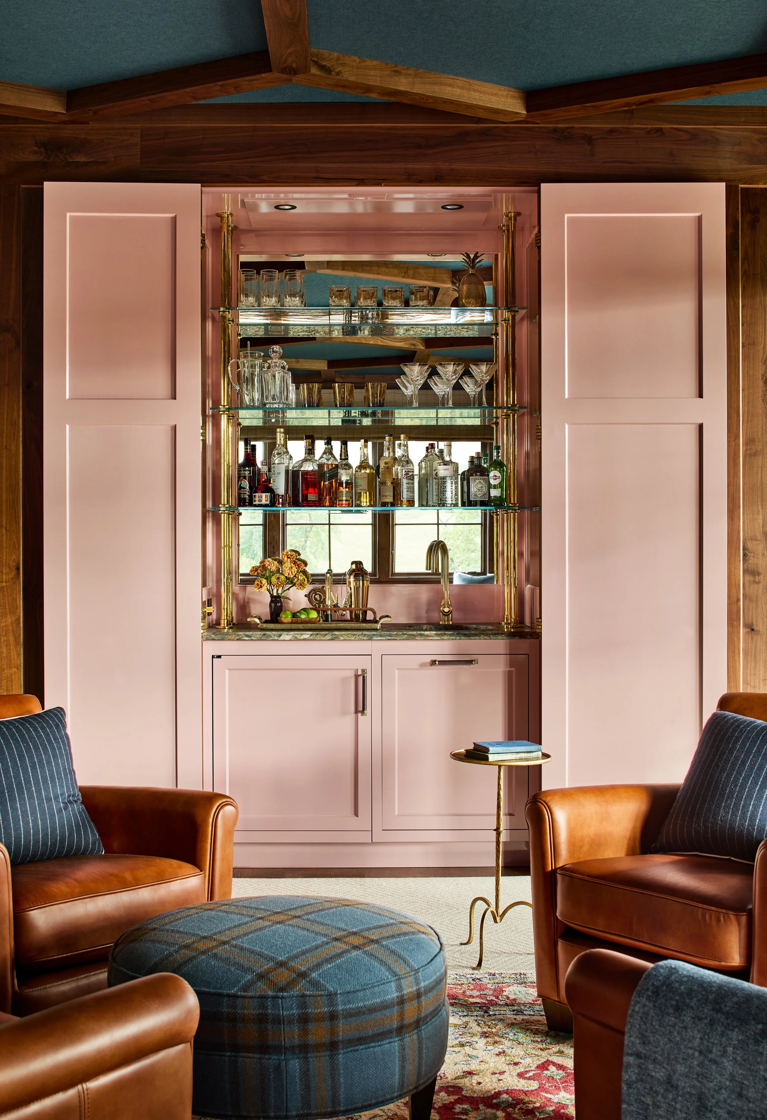 A pink built-in bar cabinet with glass shelves, holding various liquor bottles, glasses, a pineapple, and a small vase with flowers. In front, two brown leather armchairs with striped pillows, a round ottoman with plaid fabric, and a small side table