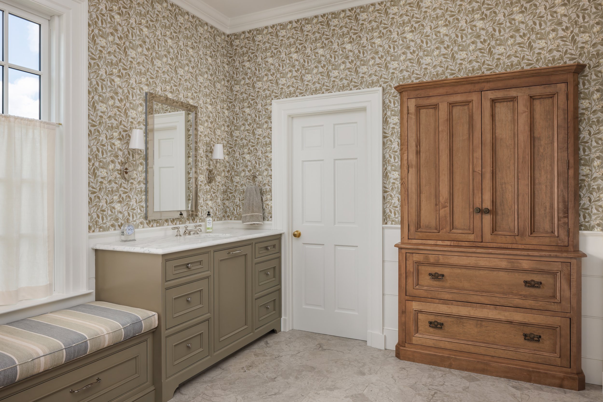 A vintage-style bathroom featuring a beige and white vanity with a marble countertop, a rectangular mirror, wall-mounted sconces, a window with a curtain, a tall wooden cabinet, and floral wallpaper.