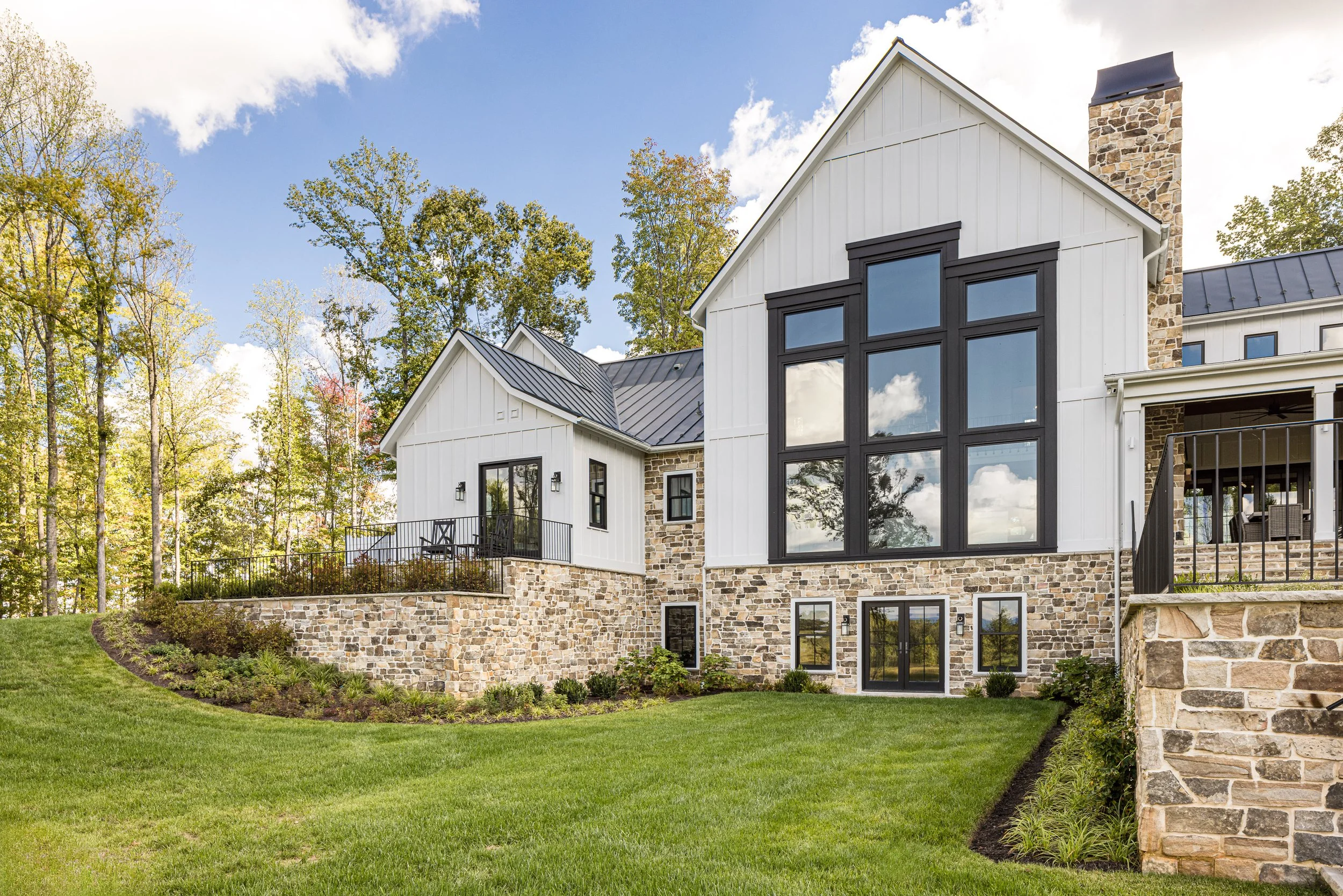 A modern house with white siding, large black-framed windows, and stone accents, situated on a grassy landscape with trees in the background under a partly cloudy sky.
