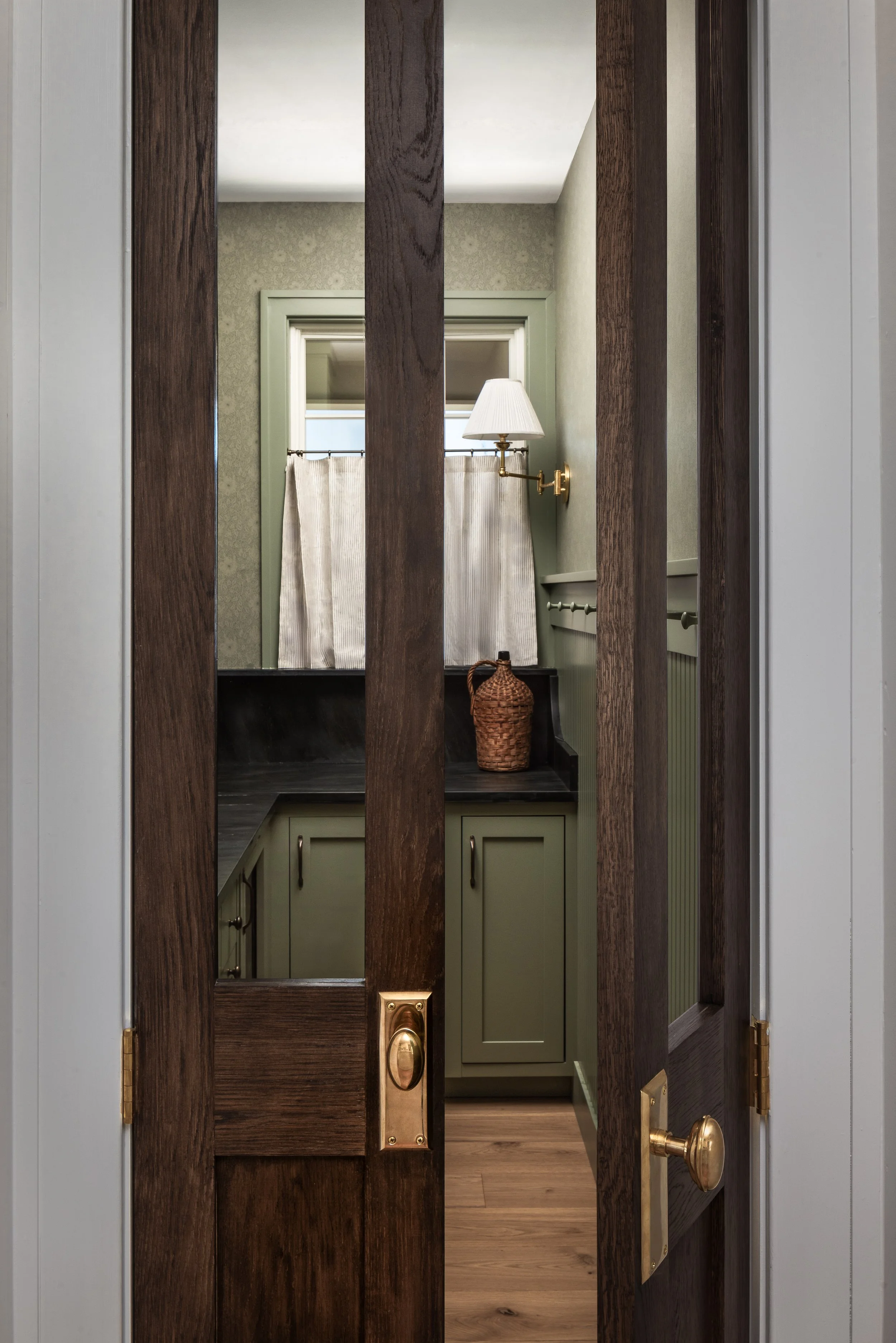 View through a partially opened wooden door reveals a vintage-style bathroom with green cabinets, a white curtain, and a wall-mounted lamp.