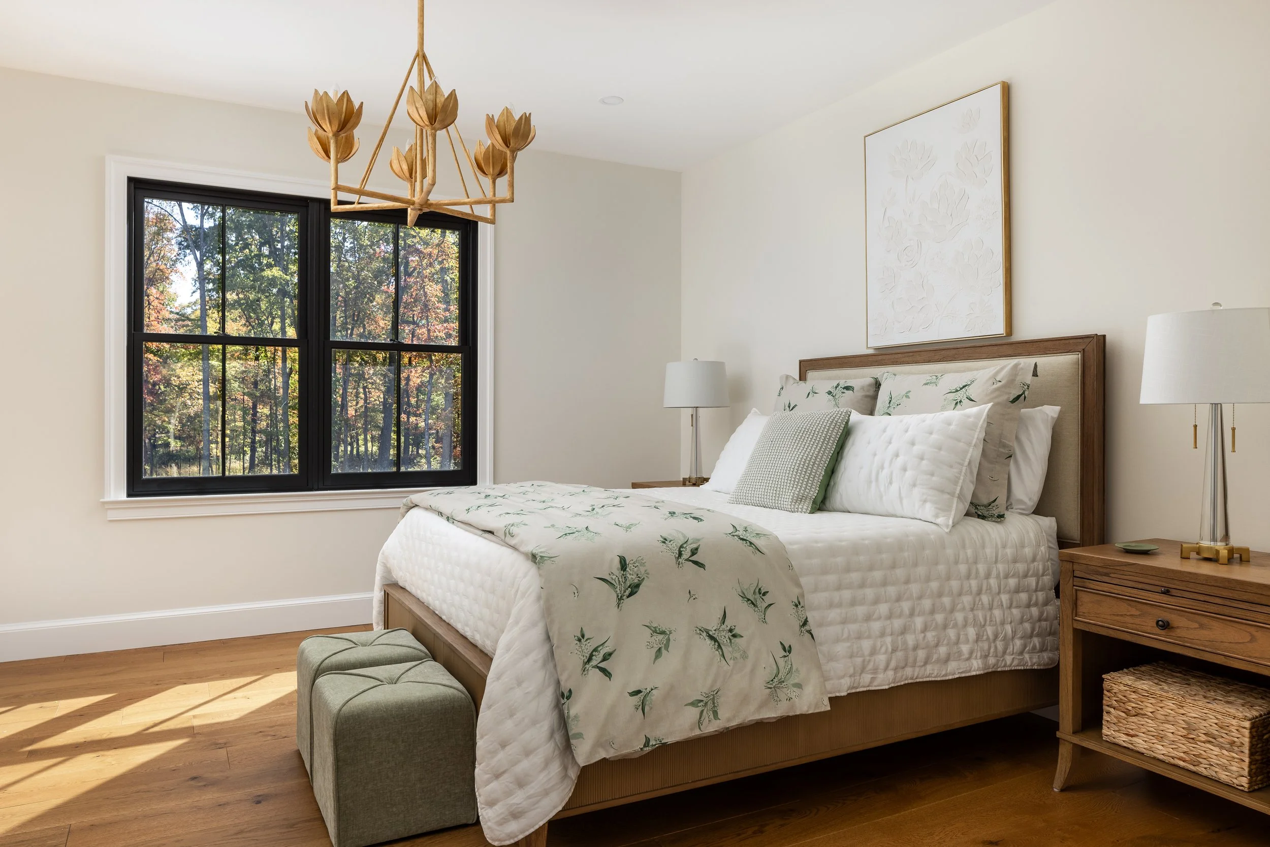 A bedroom with a large black-framed window showing trees outside. The bed has a beige headboard, white bedding with a floral pattern, multiple pillows, and a green accent pillow. There are two wooden nightstands with lamps, and a wooden bench at the 