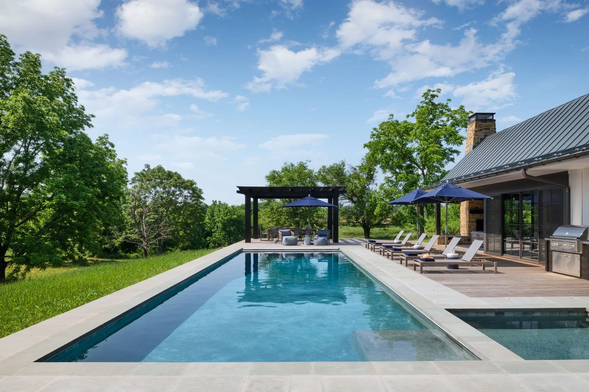 A luxurious backyard with a rectangular swimming pool, surrounded by a wooden deck with several lounge chairs and umbrellas, adjacent to a modern house with a chimney and large glass doors, lush green trees, and a partly cloudy sky overhead.