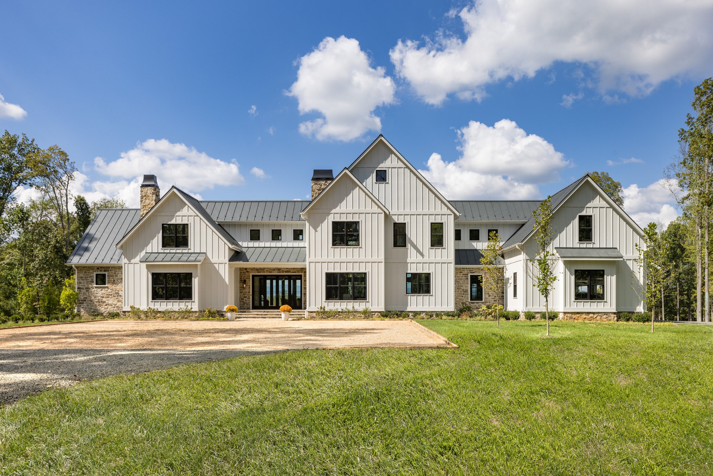 Large modern farmhouse with white vertical siding, black window frames, and metal roof, surrounded by green lawn and trees, under a partly cloudy blue sky.
