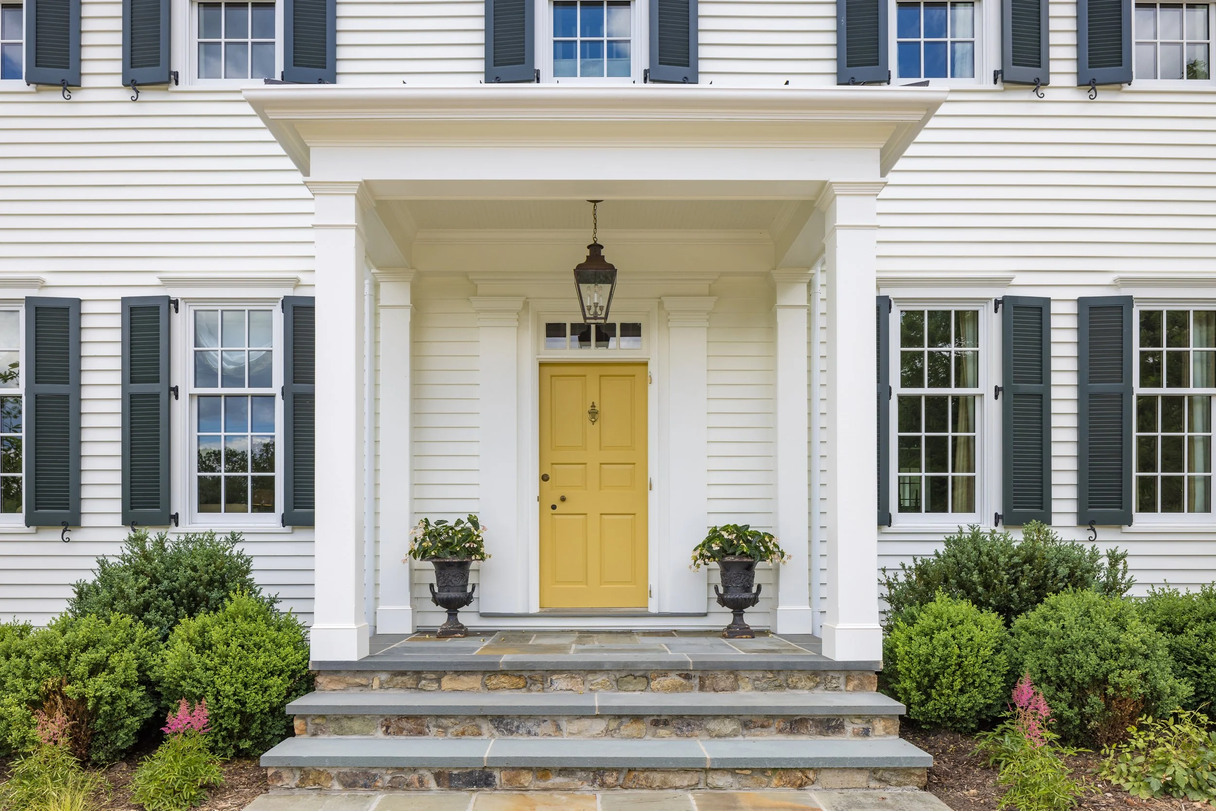 Front entrance of a white house with black shutters, a yellow door, and stone steps, decorated with potted plants.