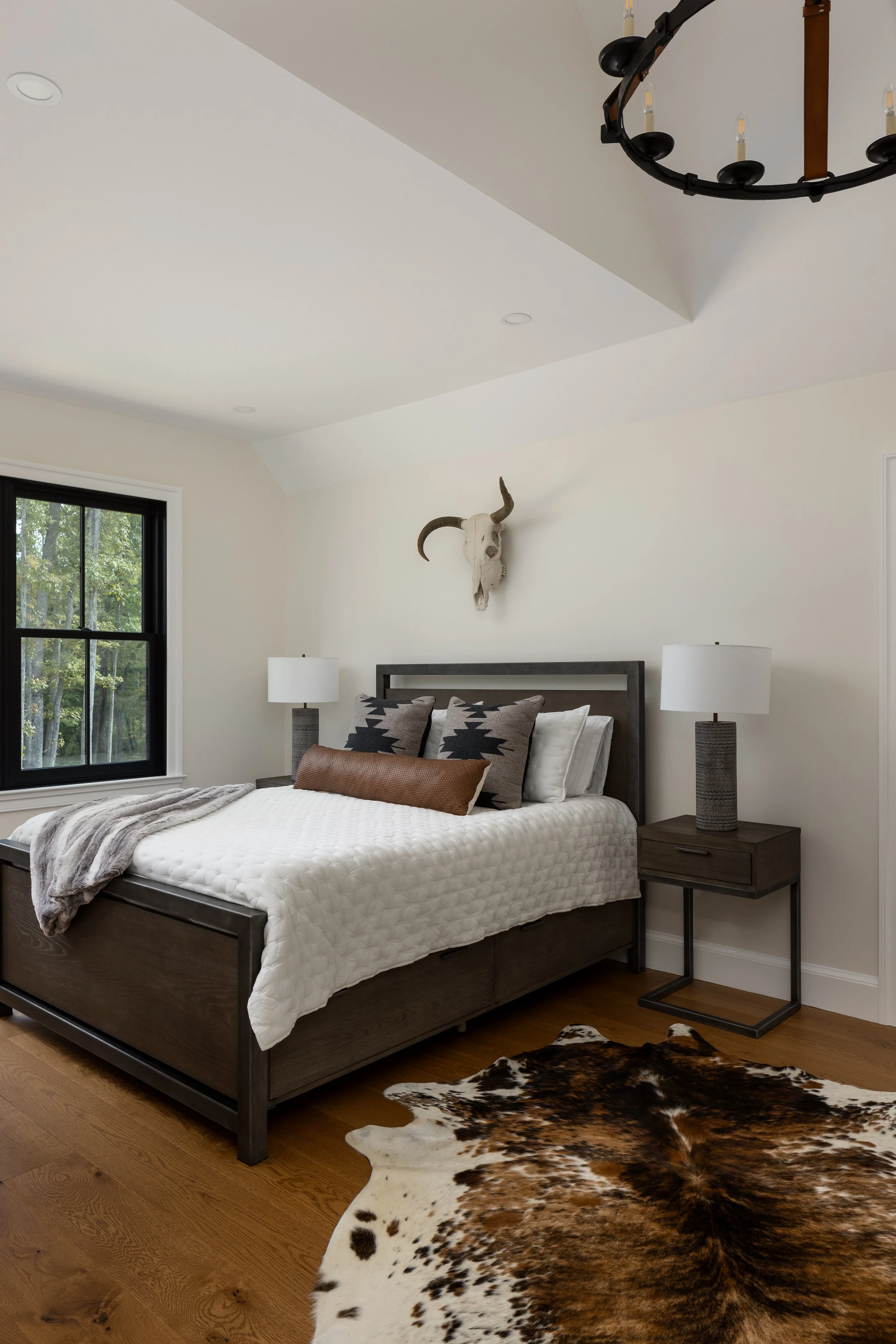 Bedroom featuring a dark wooden bed with white quilted bedding, decorative pillows, and a faux fur throw. On the wall behind, a mounted animal skull with horns. Two matching bedside tables with white lamps. A cowhide rug on the wooden floor. A window