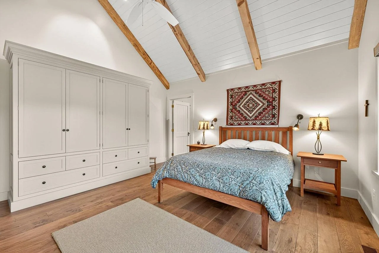 A bedroom with a wooden bed frame, blue patterned bedspread, two nightstands with lamps, a large white wardrobe, a hanging textile art, and a vaulted ceiling with wooden beams.