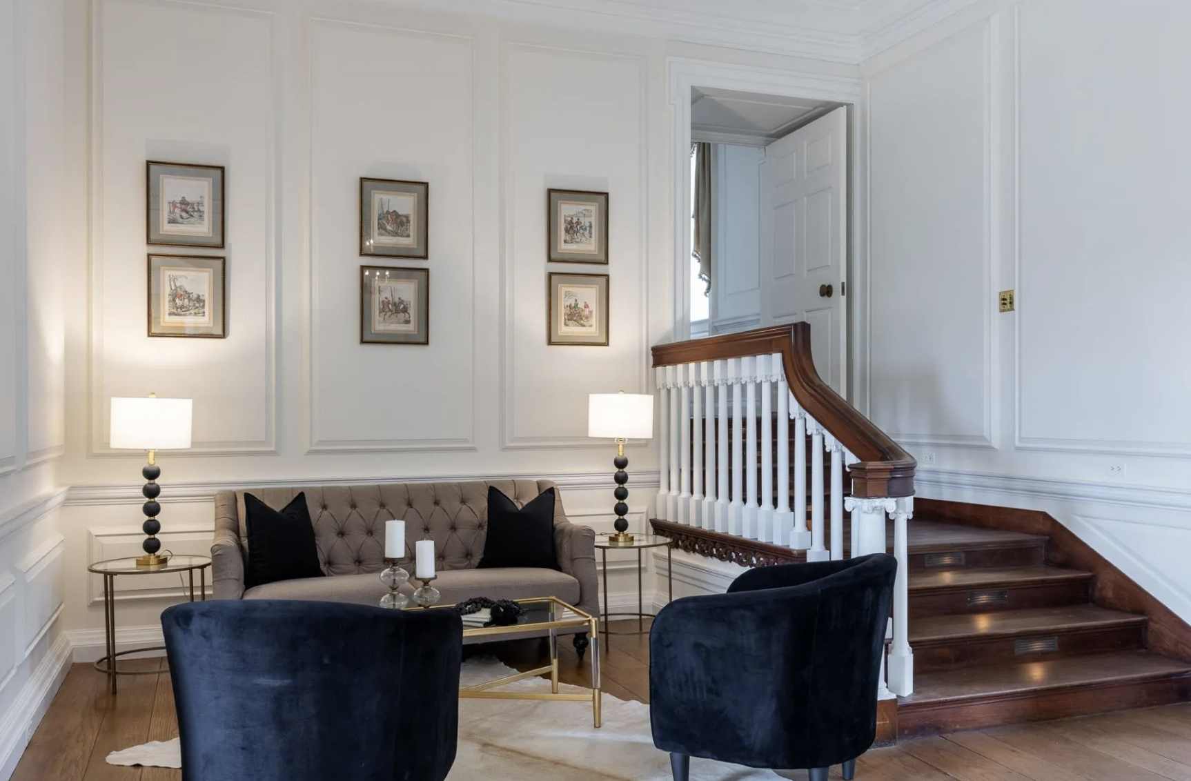 Elegant living room with white paneled walls, a beige tufted sofa with black pillows, two gold side tables with lamps, a black velvet armchair, a glass coffee table, and a staircase with wooden steps and white balustrade.