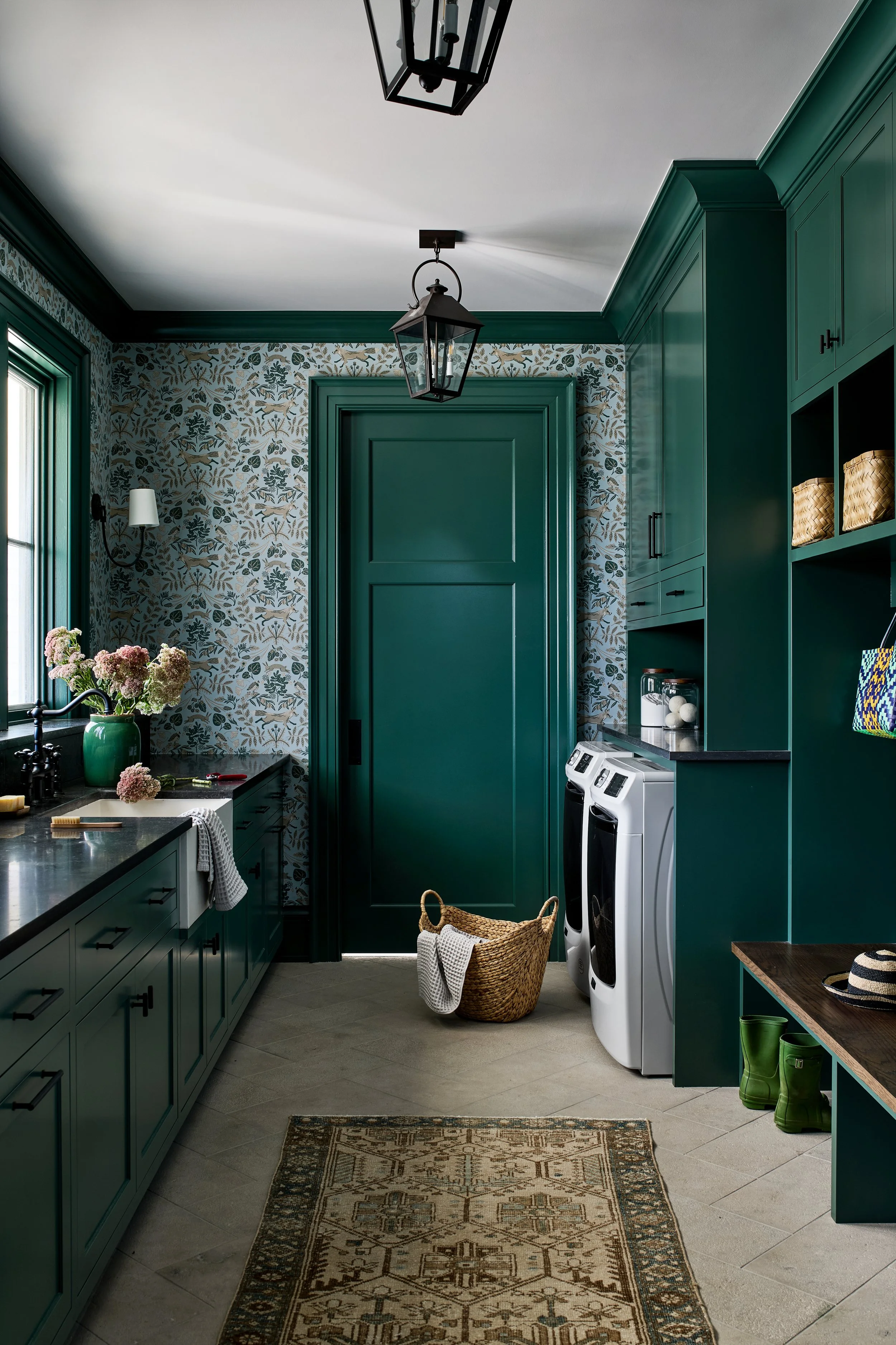 A laundry room with dark green cabinets, a window with a bouquet of pink flowers, a washer and dryer, and a basket with a towel. The room has patterned wallpaper, a rug, and some green rain boots.