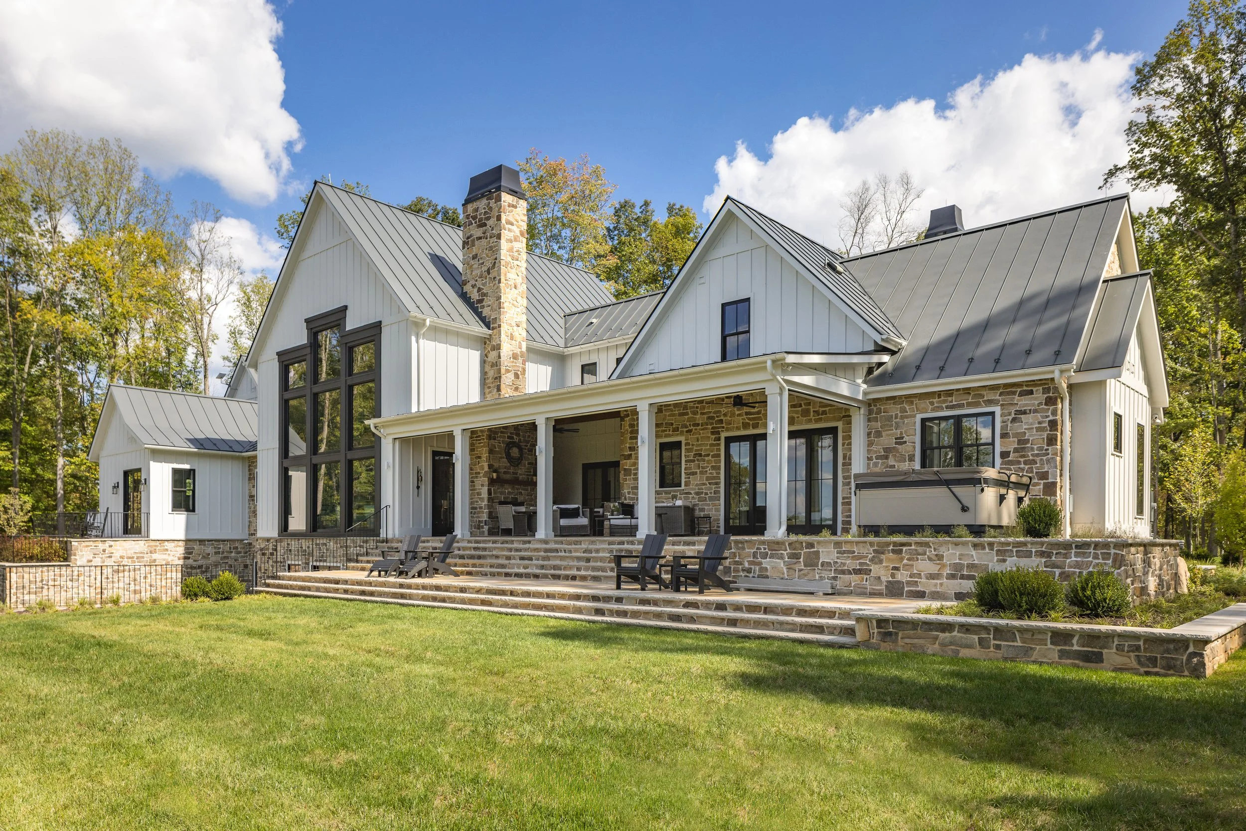 Modern house with a stone and white exterior, large windows, a metal roof, a chimney, front steps, outdoor seating area, and lush green lawn surrounded by trees.