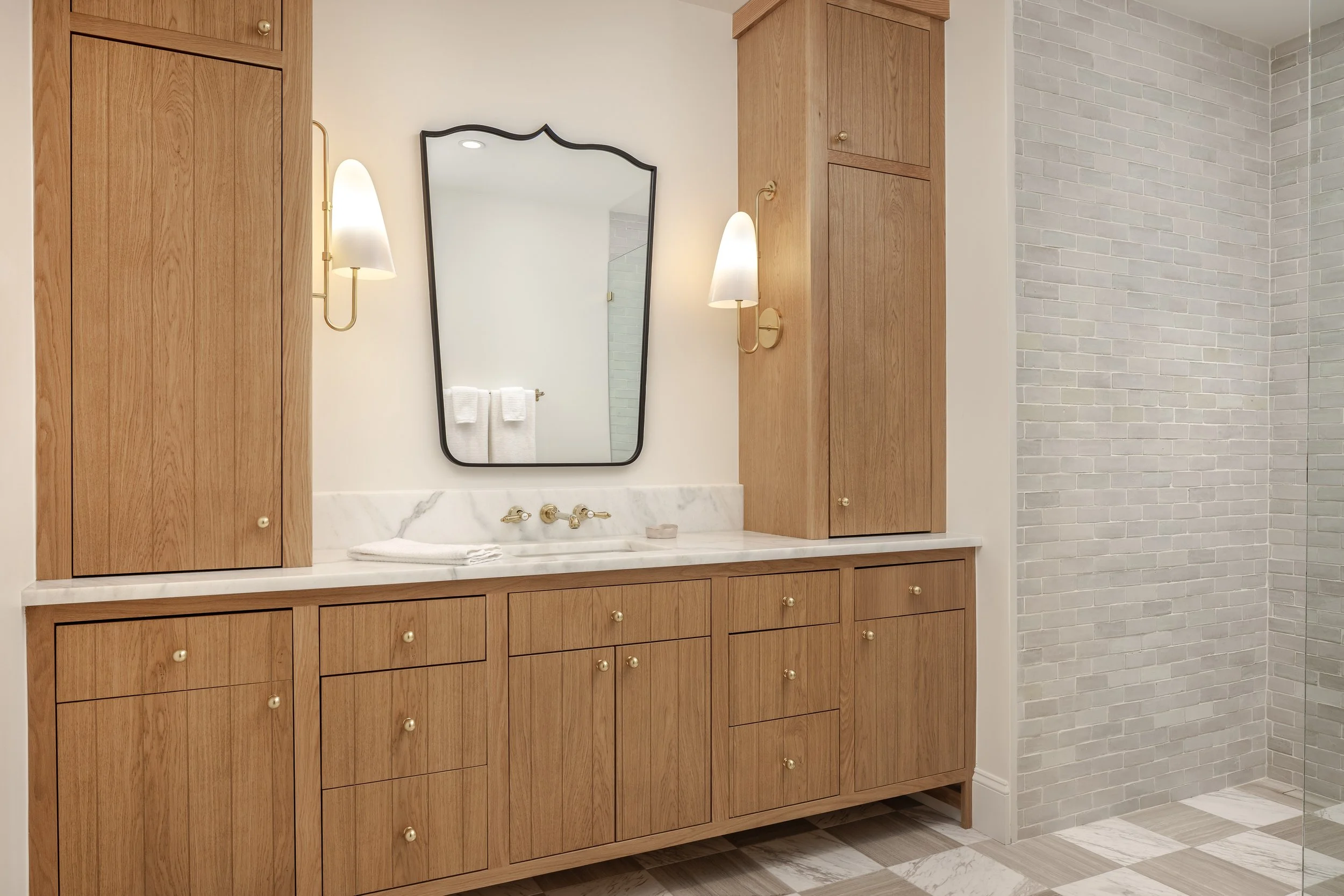 Elegant bathroom vanity with wooden cabinets, a white marble countertop, brass fixtures, and a large mirror. On the right, a walk-in shower with white brick tiles and a glass enclosure.