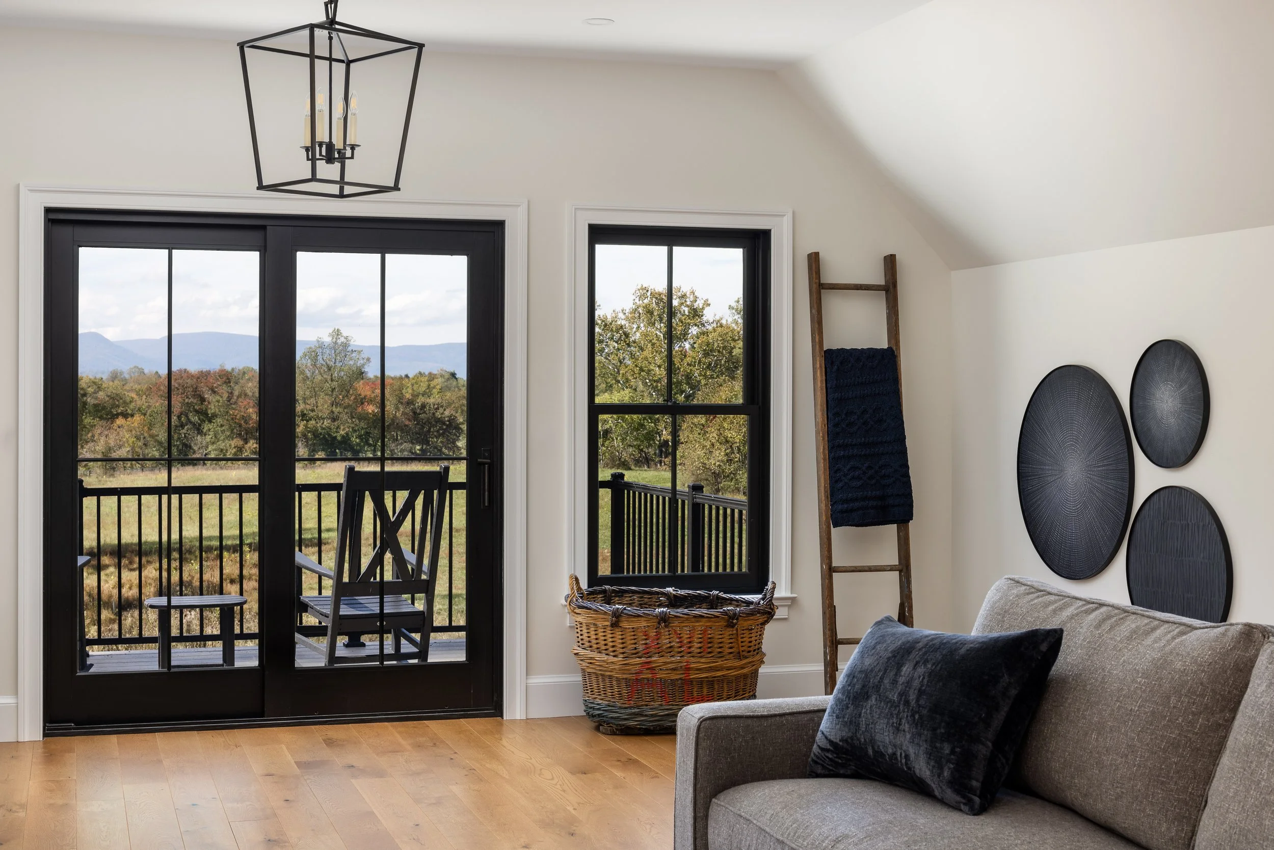 Living room with a sliding glass door leading to a balcony, a window, a wicker basket, a leaning wooden ladder with dark blanket, black wall art, and a beige sofa with black pillows.