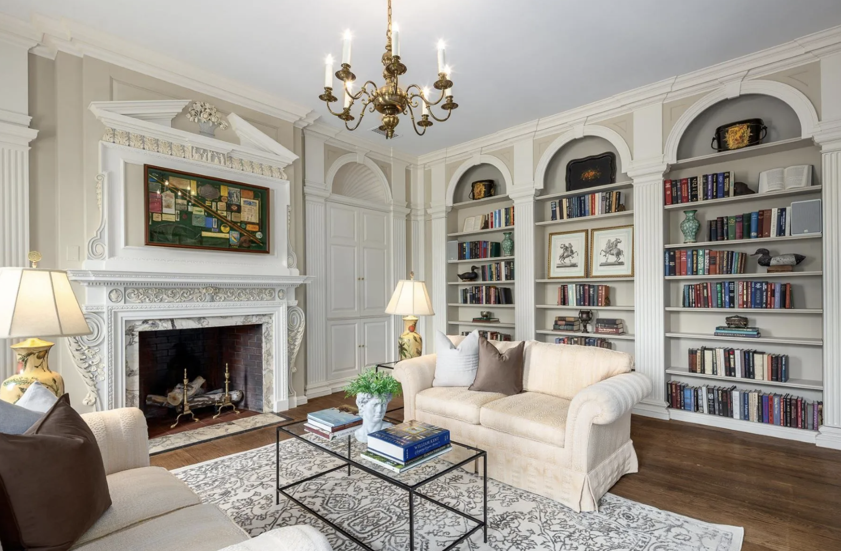 Elegant living room with white ornate fireplace, floor-to-ceiling built-in bookshelves filled with books and decor, beige sofa with cushions, and a black coffee table with books, set on a patterned area rug. A brass chandelier hangs from the ceiling,