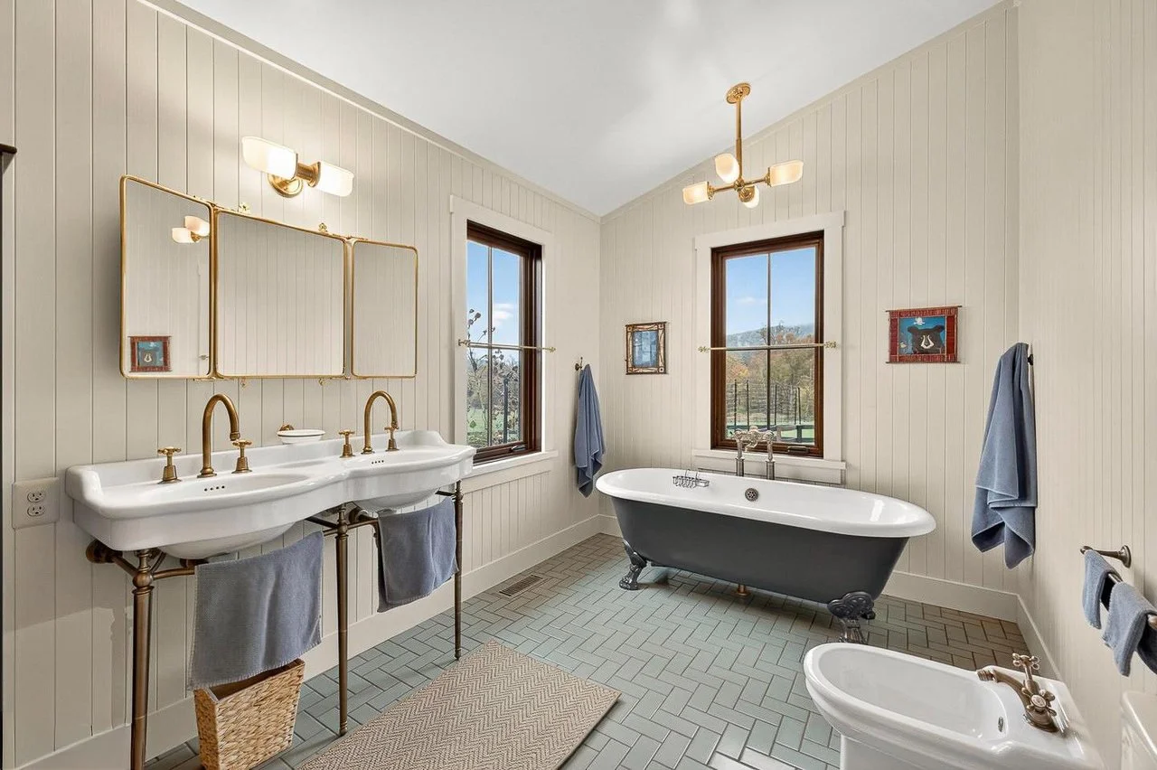 Bathroom with double sink vanity, freestanding bathtub, bidet, beige beadboard walls, wooden window frames, and vintage-style fixtures.