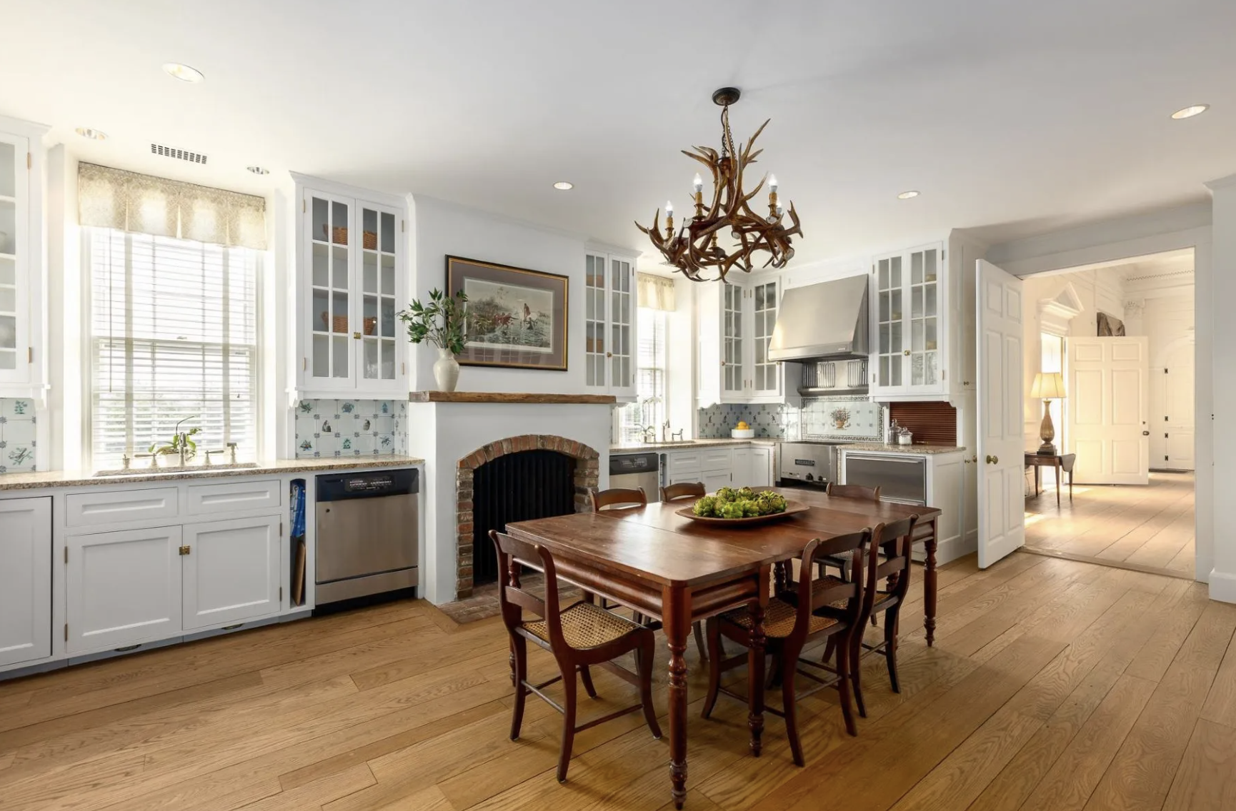 A bright kitchen with white cabinets, a wooden dining table with six chairs, a brick fireplace, and a chandelier made of antlers.