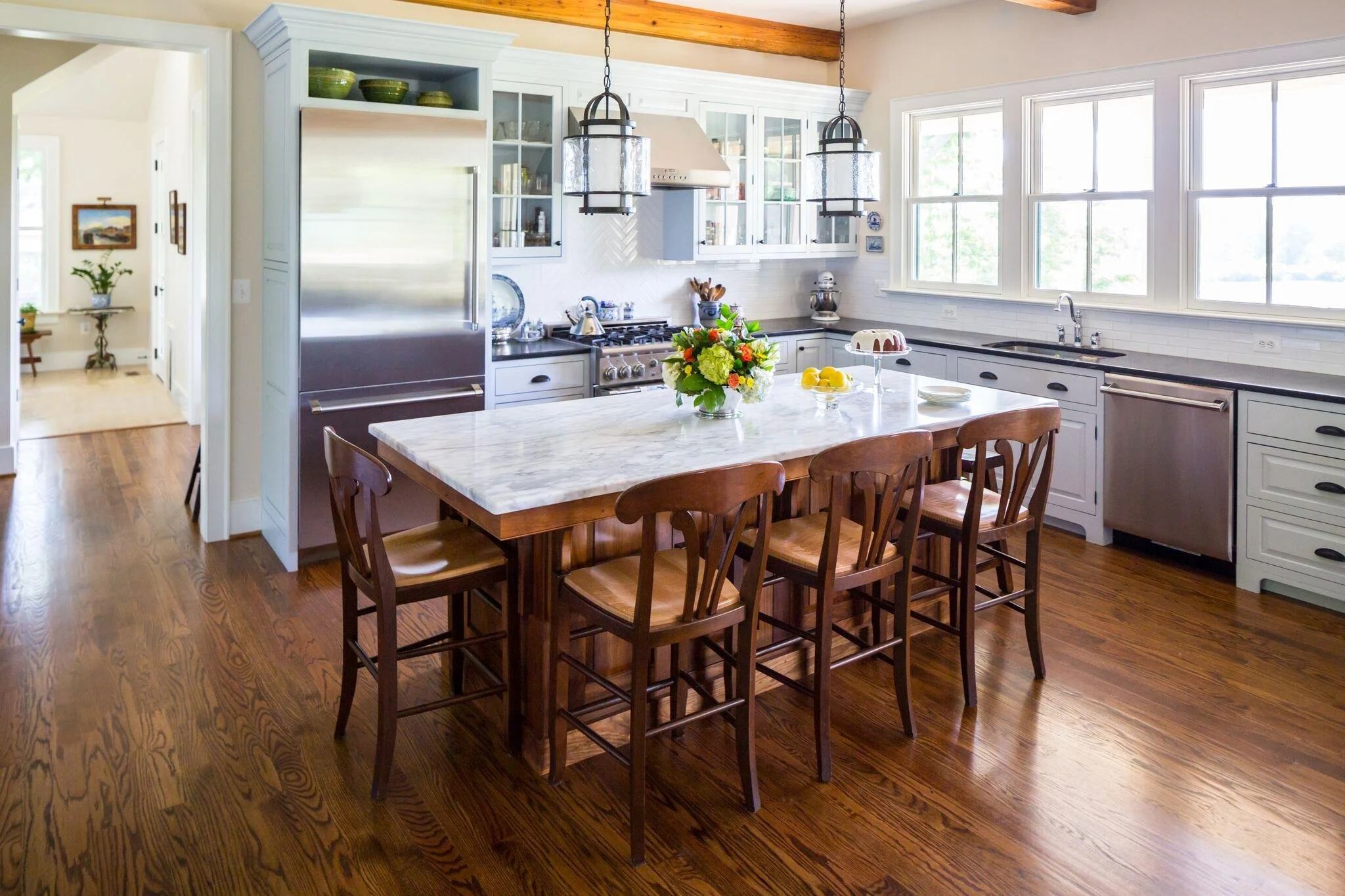 Bright kitchen with white cabinets, marble island, wooden chairs, hardwood floors, large windows, and hanging light fixtures.