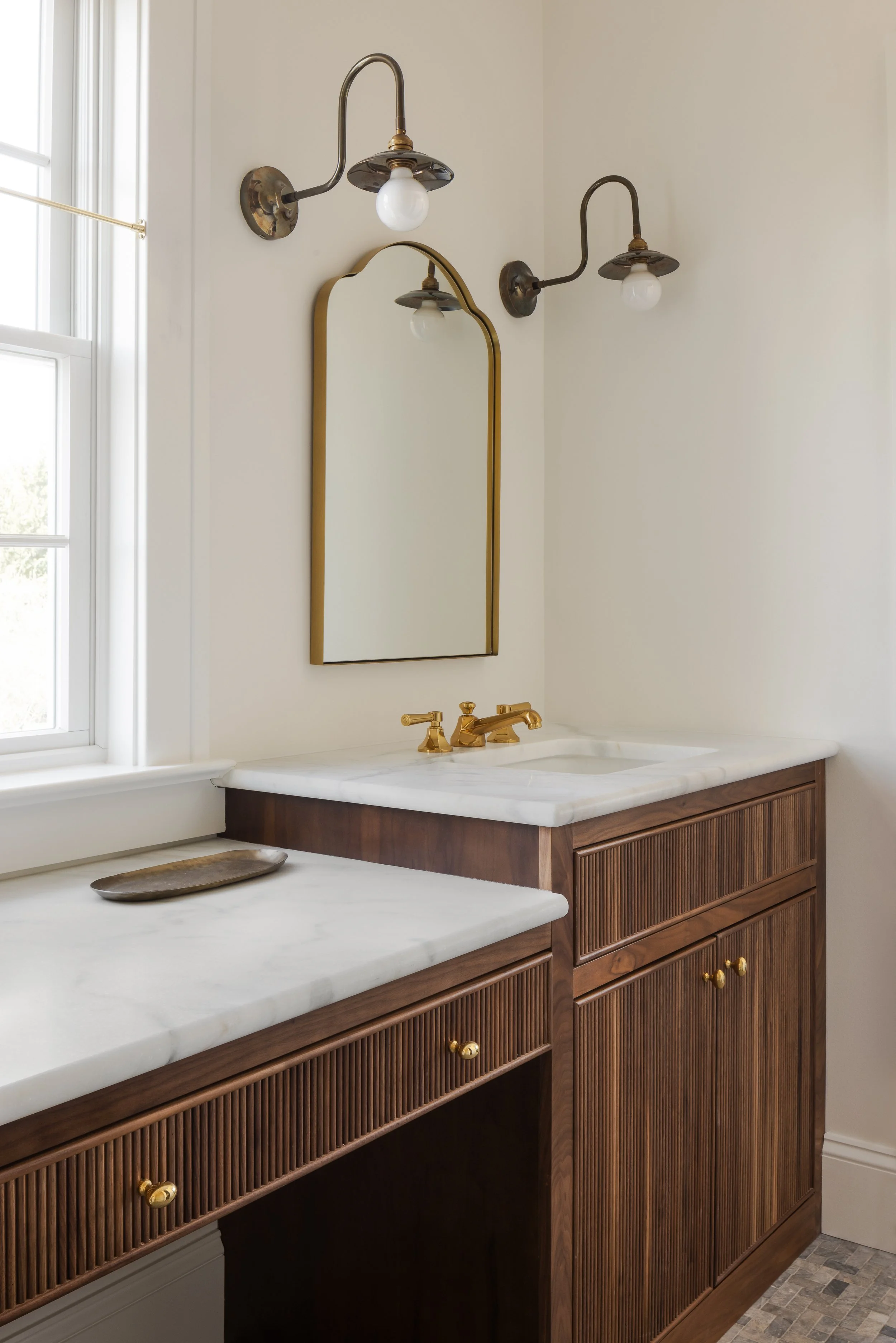 Bathroom vanity with white marble countertop, brass faucet, wooden cabinet with vertical grooves, and a gold-framed mirror. Two vintage wall sconces with exposed bulbs are above the mirror, and a window with white trim is nearby.