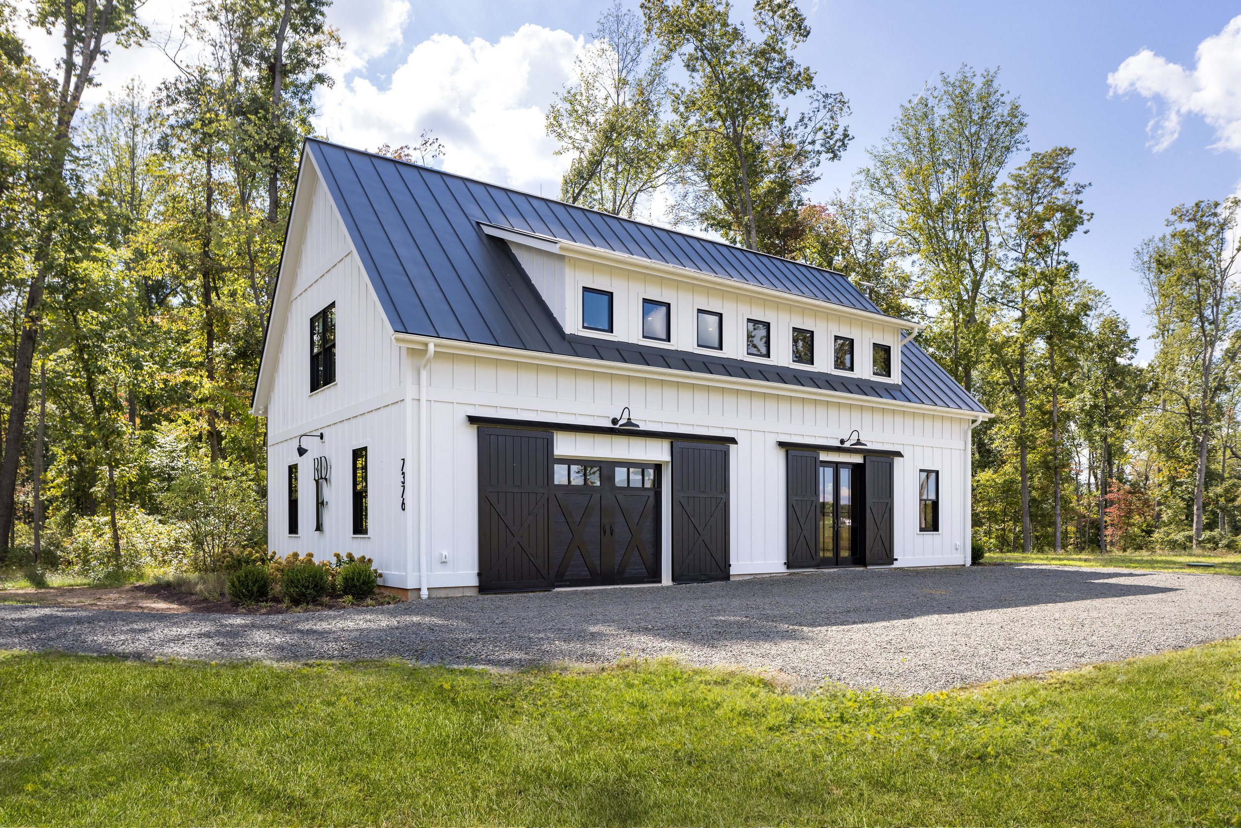 A modern white barn with black accents and a metal roof, situated on a grassy and gravel lot surrounded by trees.