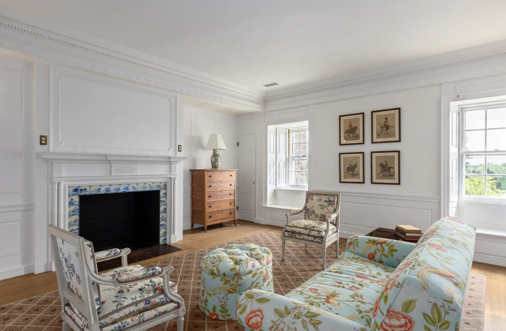 A cozy living room with white walls, a fireplace with decorative tile, floral upholstered furniture including a sofa, armchair, and ottoman, hardwood floors, a wooden dresser with a lamp, and framed horse artwork on the walls.