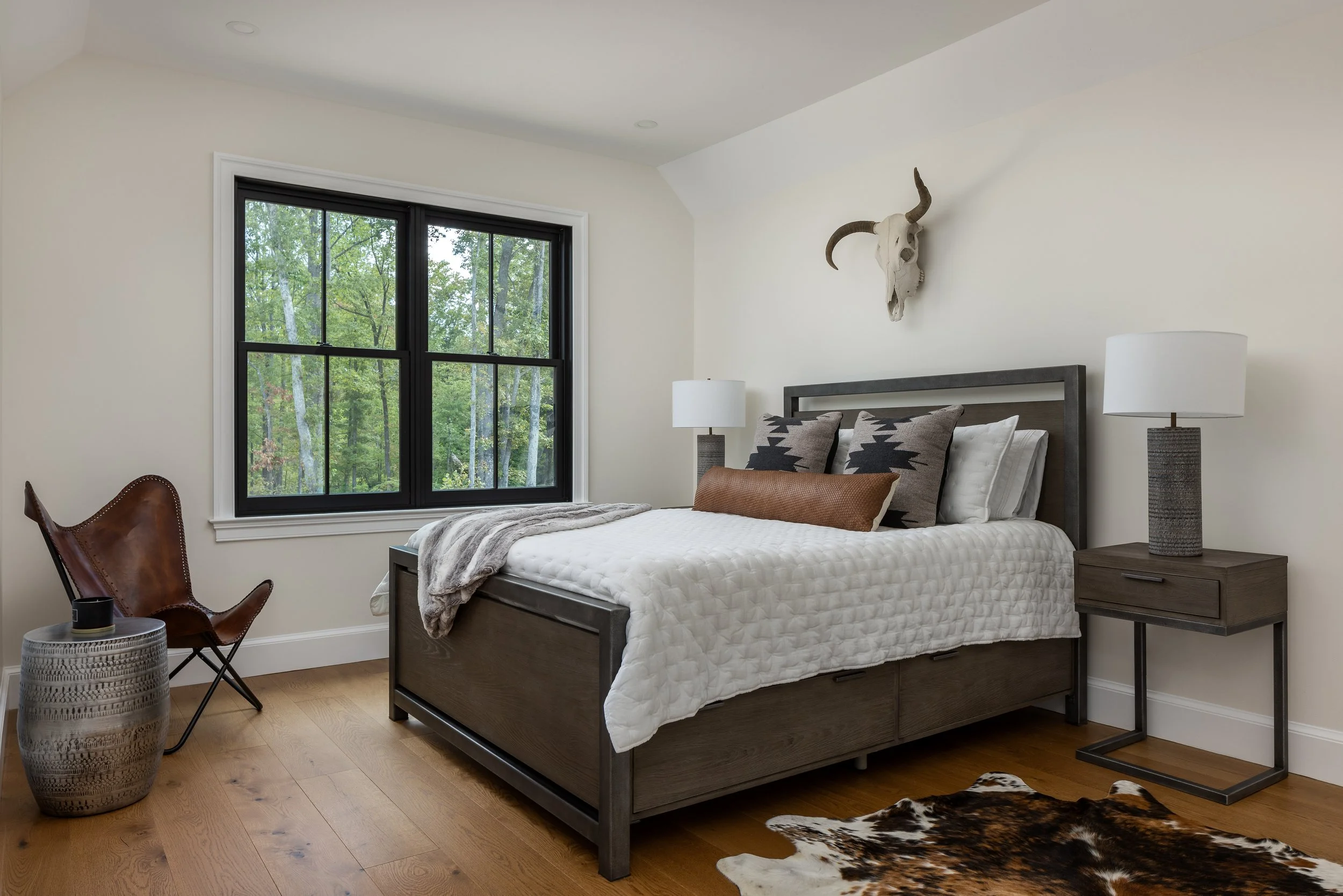 Modern bedroom with large window, wooden floor, bed with white and patterned pillows, gray and brown furniture, and animal skull decor on the wall.