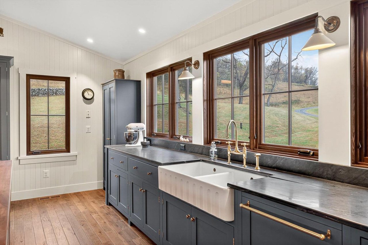 Kitchen with gray cabinets, white farmhouse sink, large windows showcasing a green outdoor landscape, hardwood floor, and brass fixtures.