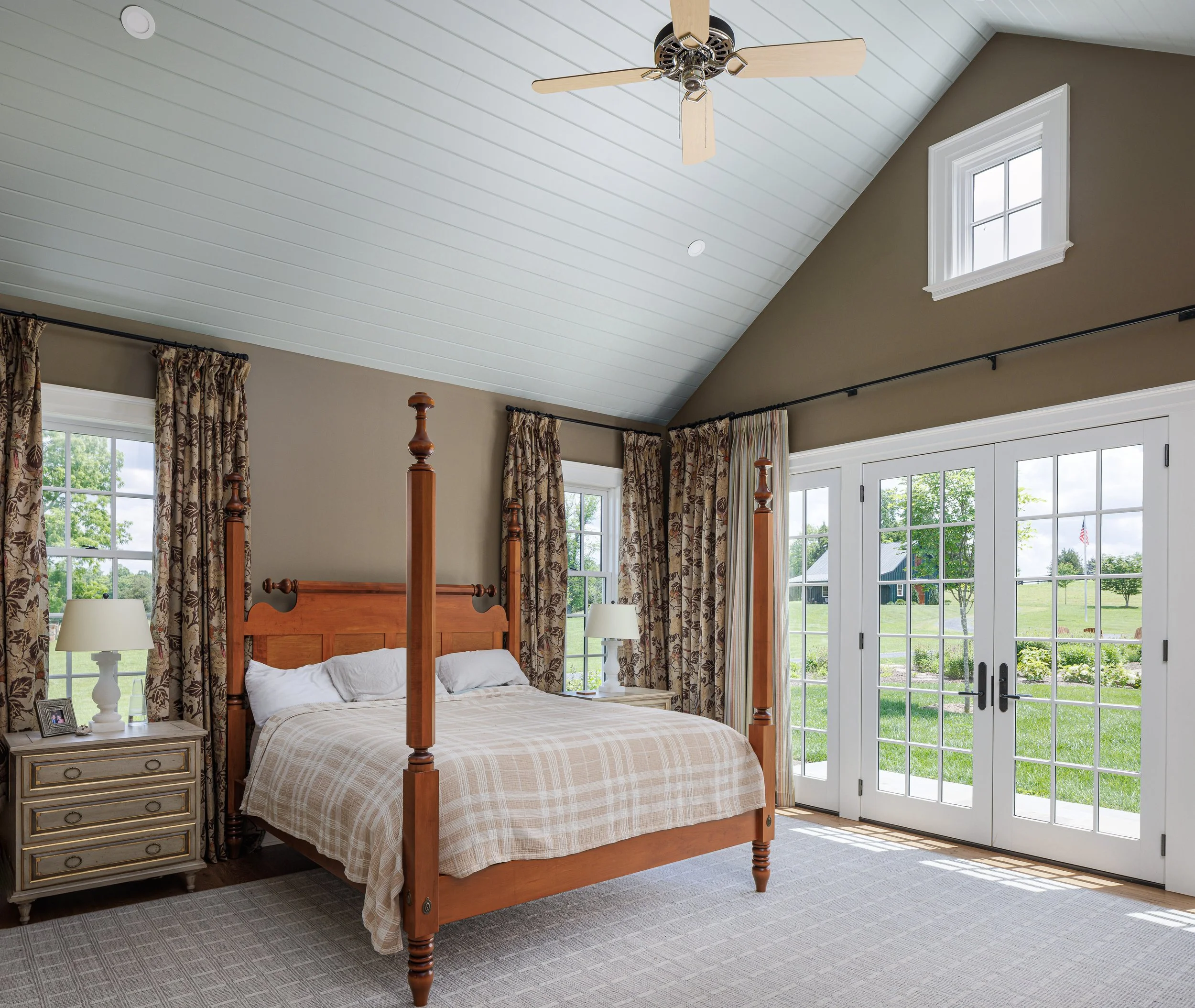 A bedroom with a four-poster bed, nightstands, lamps, patterned curtains, and large French doors leading to a green outdoor landscape, with a small window near the ceiling.