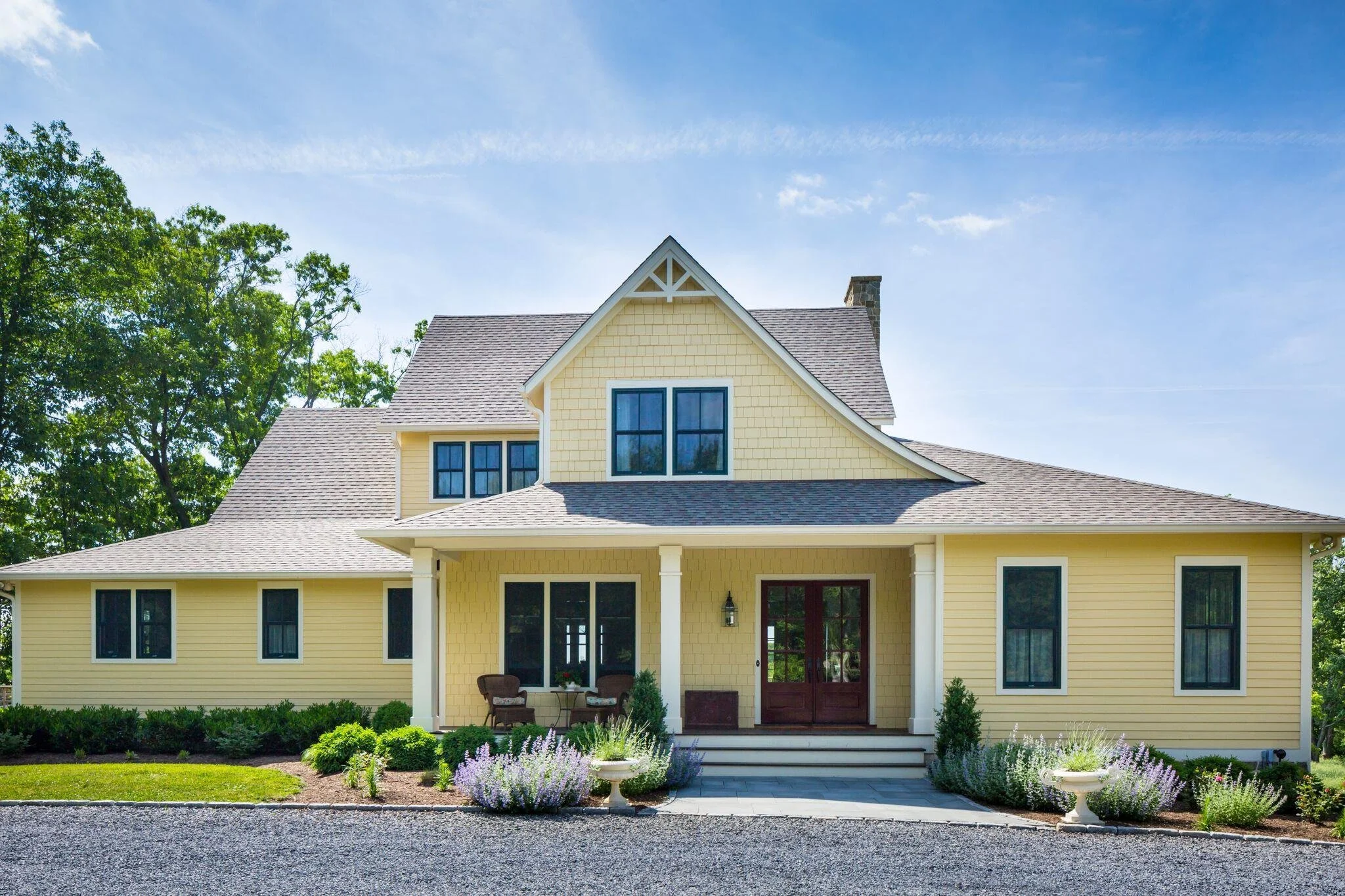 A yellow house with a covered porch, front steps, and black window frames, surrounded by manicured shrubs and flowers under a blue sky.