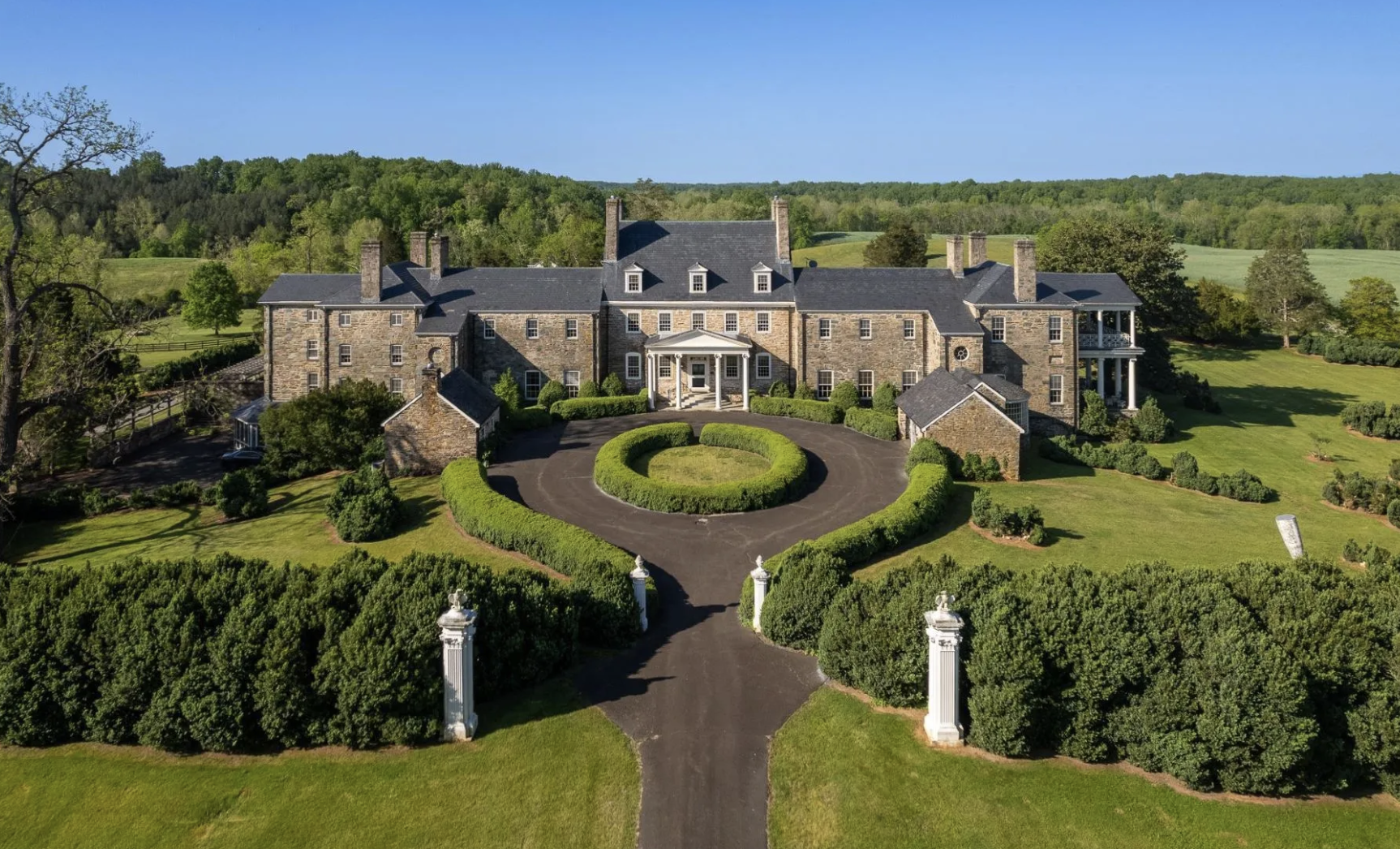 A large stone mansion with multiple chimneys, surrounded by well-manicured gardens and lush green lawns, with a circular driveway and decorative white pillars at the entrance, set against a backdrop of rolling hills and trees.