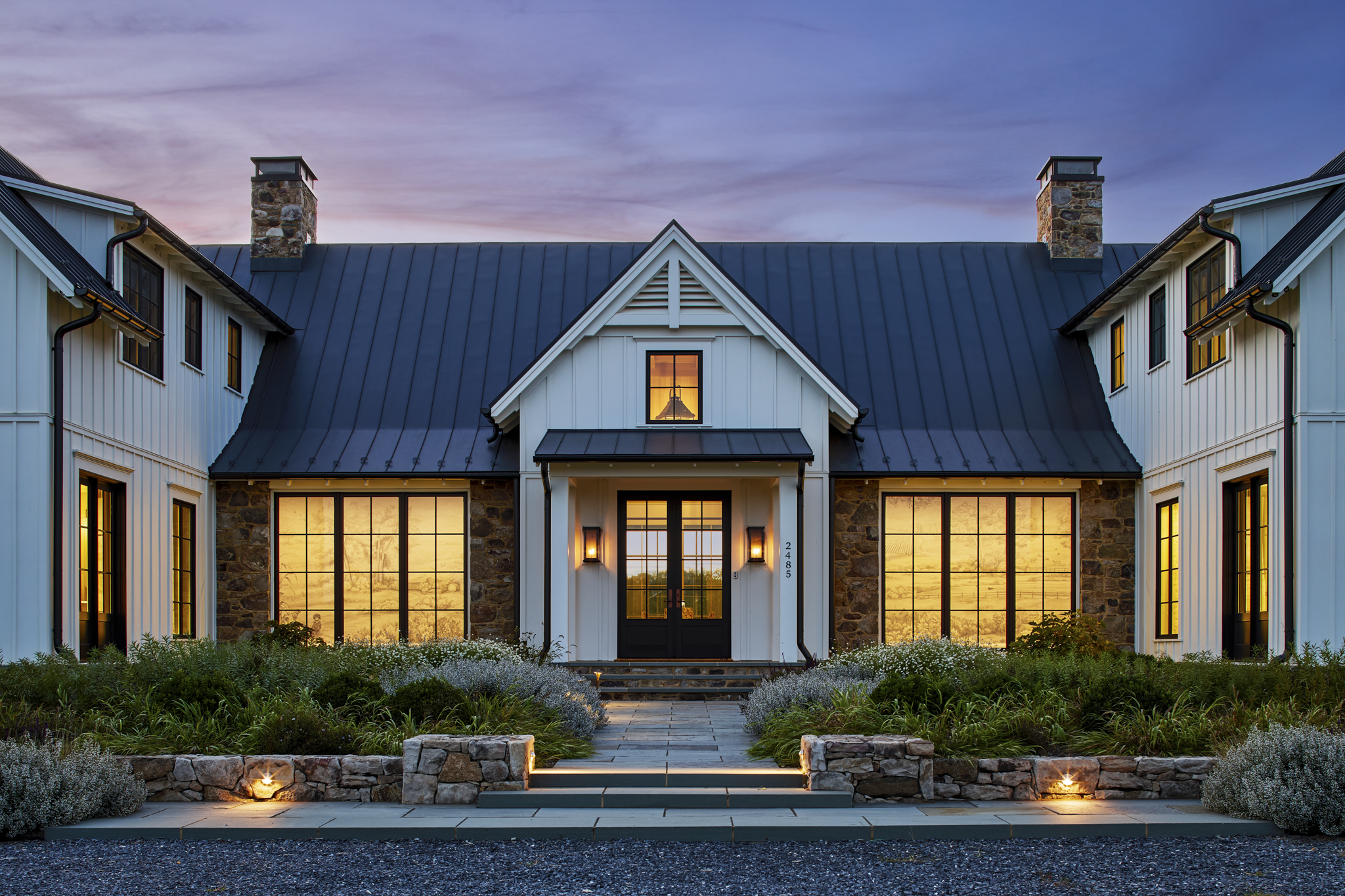 A modern two-story house with a stone and white siding exterior, large windows, and a black front door. It is illuminated from inside, with a sunset sky in the background.