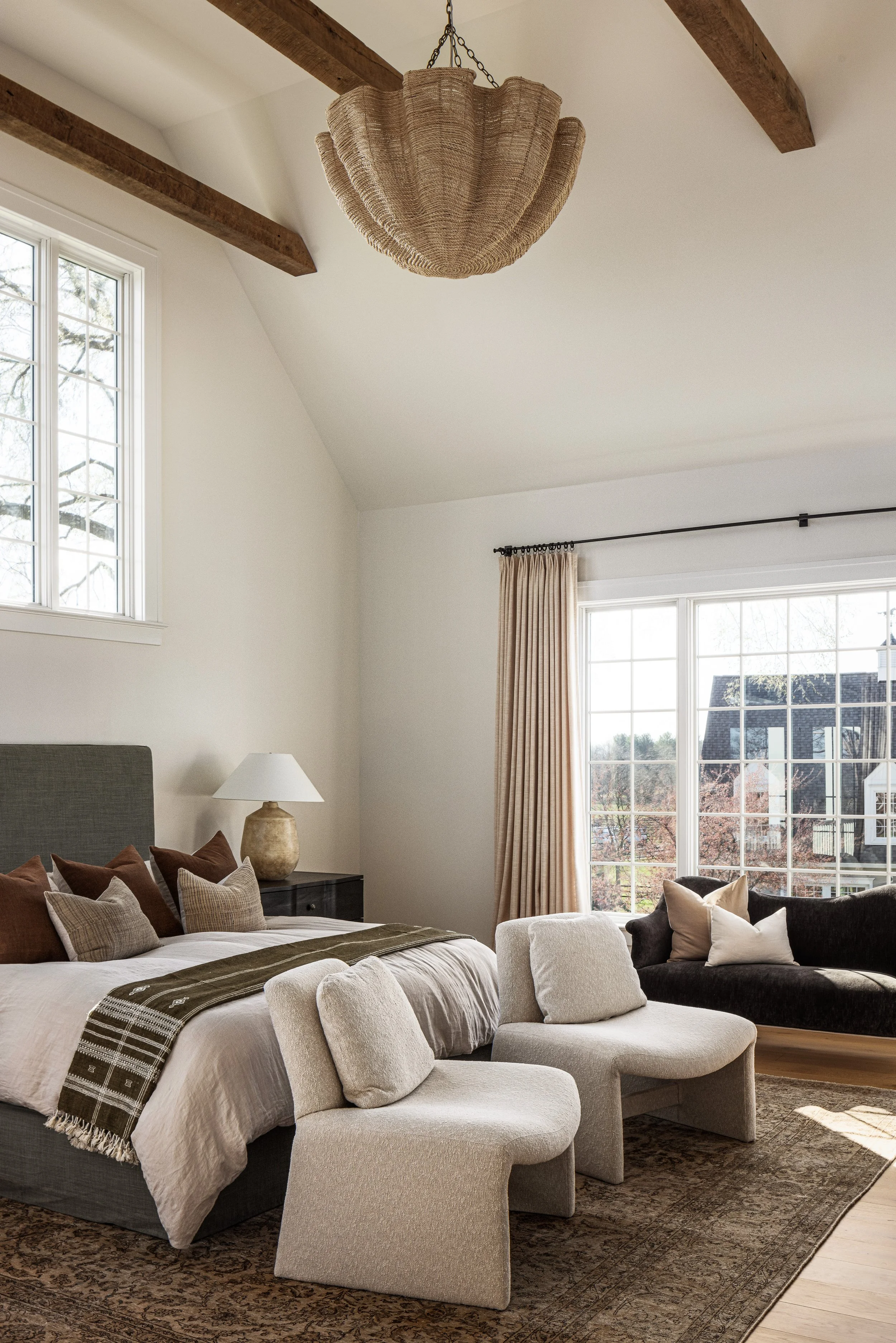 A bedroom with a large bed, beige and brown pillows, a textured beige blanket, a nightstand with a lamp, and two white upholstered chairs. There are large windows with curtains, a dark sofa with pillows, and wooden ceiling beams with a woven pendant 