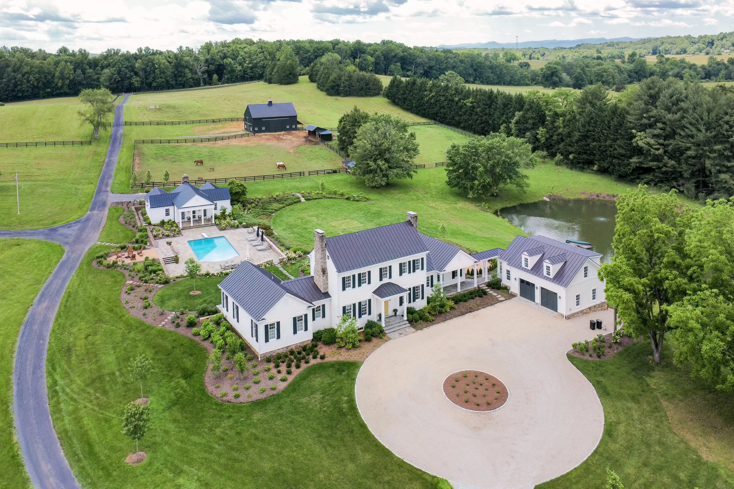 A large white estate with a circular driveway, pool, detached garage, surrounded by green lawns, trees, a pond, and farmland fenced for horses.