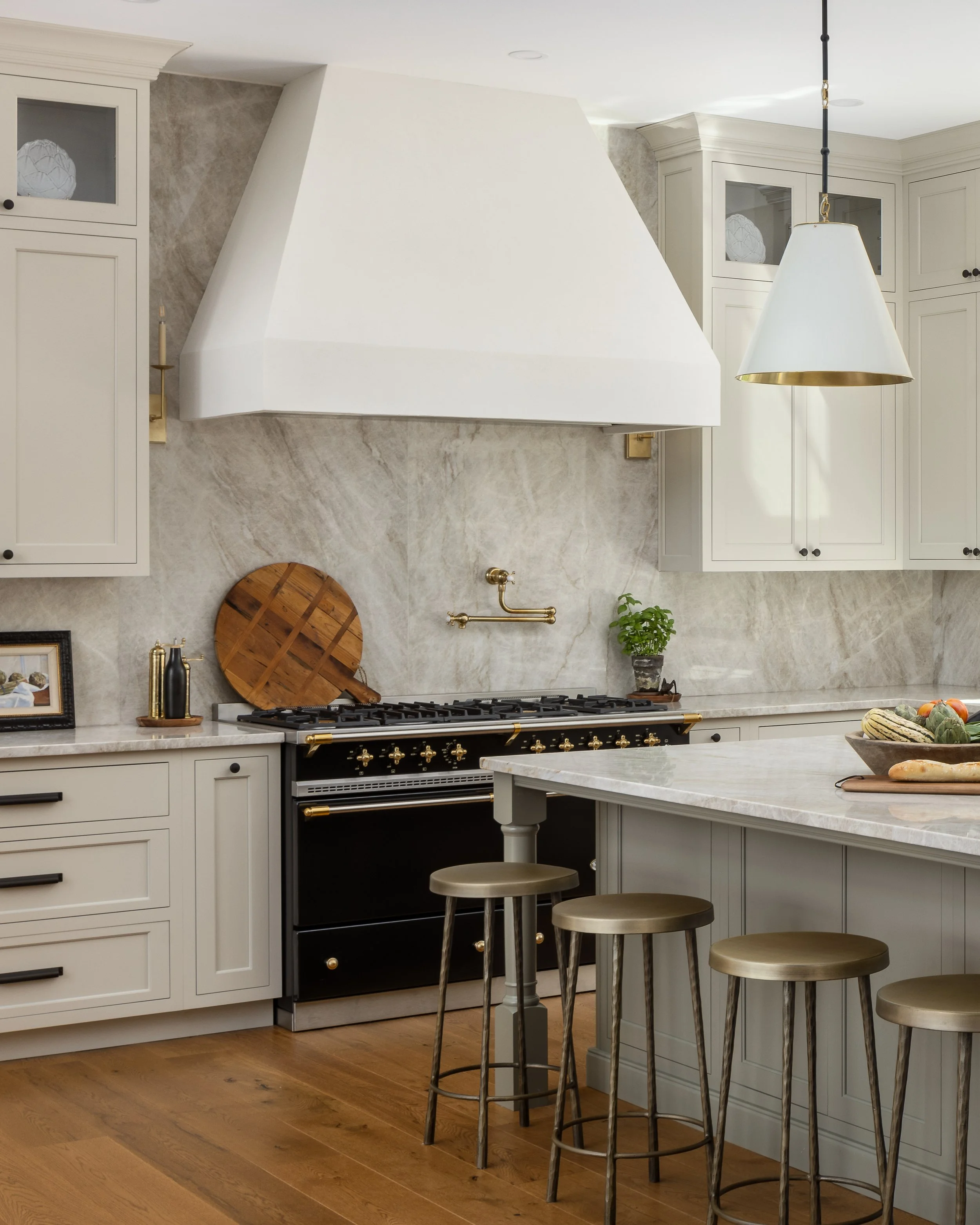 Modern kitchen with white cabinetry, a black oven with gold details, a marble backsplash, a large white vent hood, a wooden cutting board, and a kitchen island with three metallic barstools.