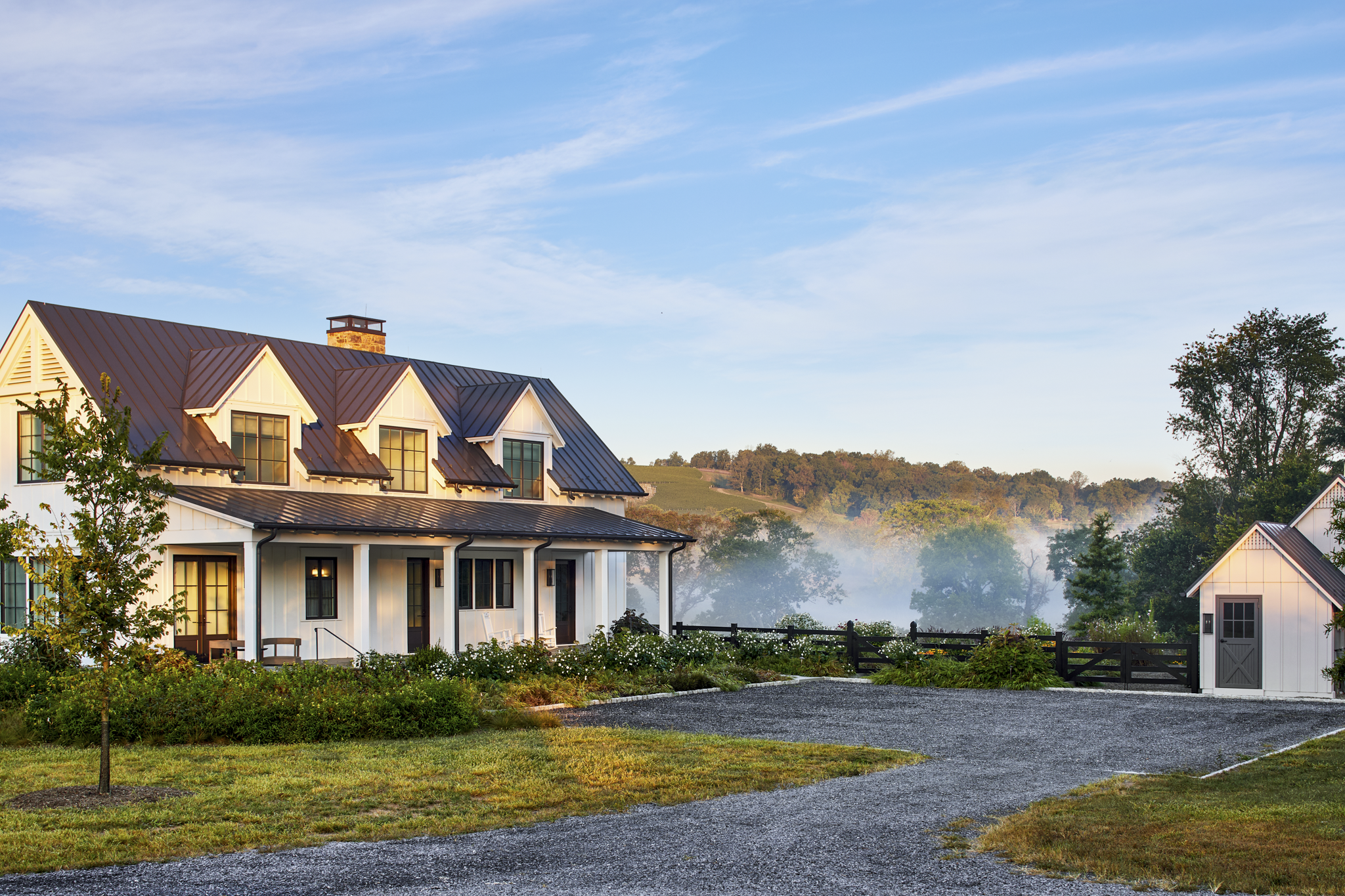 A large white house with a dark metal roof, multiple front-facing windows, and a chimney. There is a gravel driveway, green lawn, and some small trees, with a scenic background of rolling hills and a partly cloudy sky.