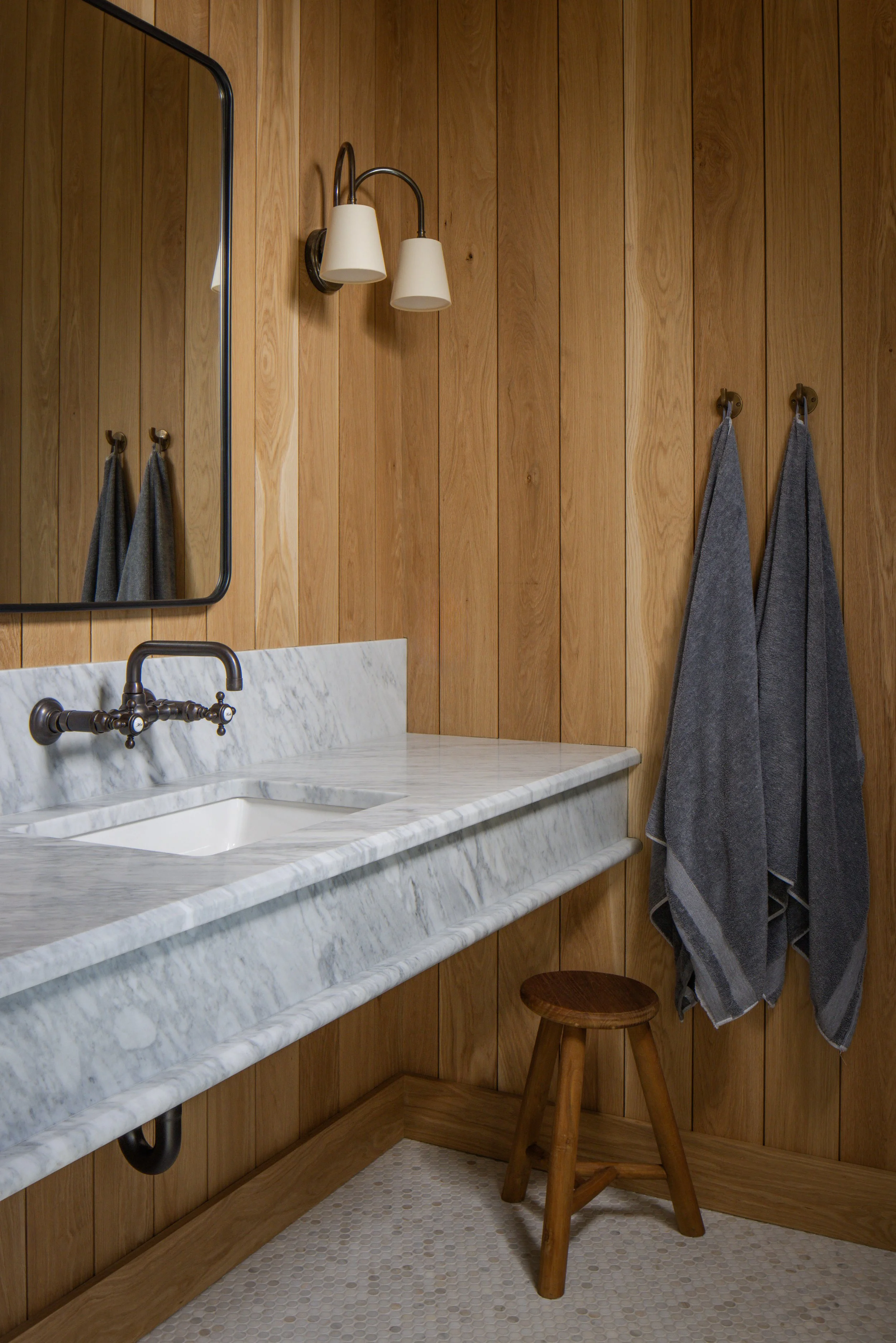 Bathroom with wooden panel walls, marble countertop with a sink, wall-mounted faucet, mirror, wall lamp with two shades, and two gray towels hanging on hooks beside a wooden stool.