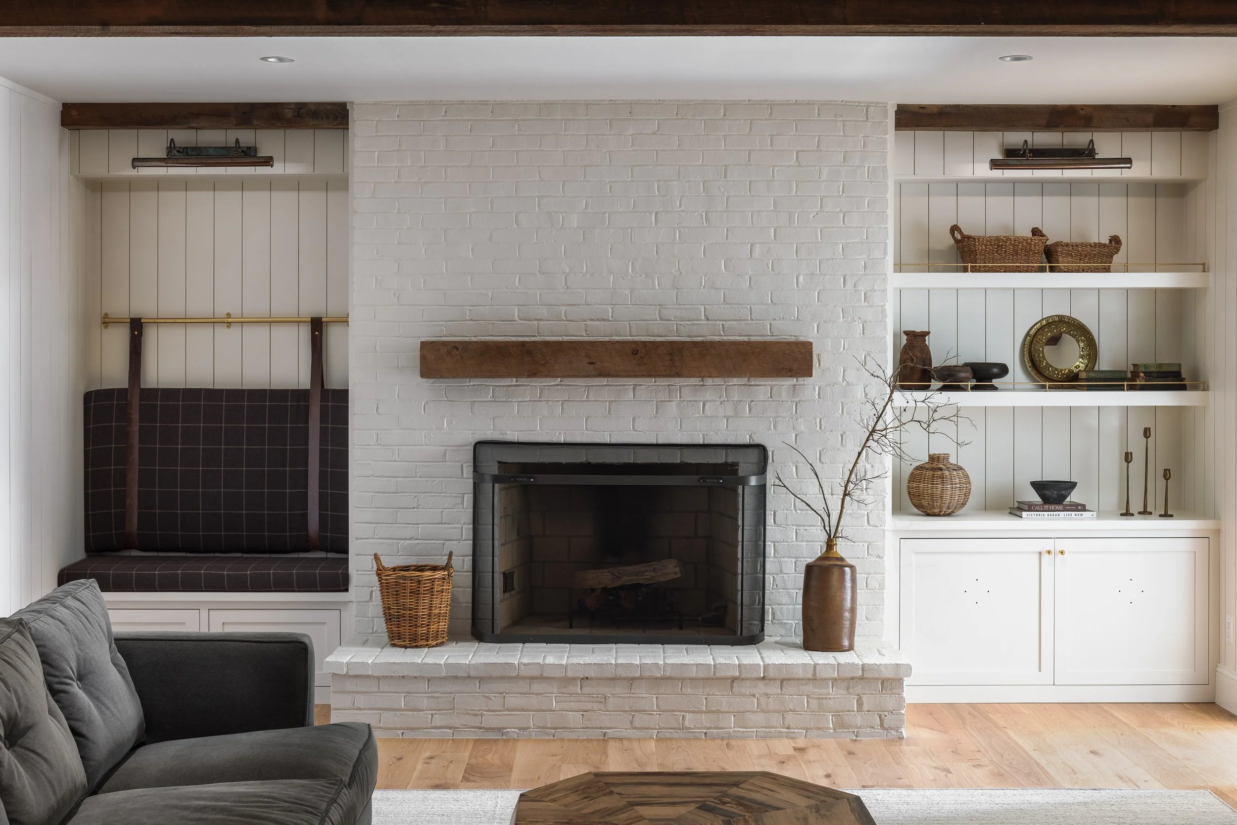 Living room with white brick fireplace, wooden mantel, built-in shelves with decor, gray couch, hardwood floors, and beige walls.