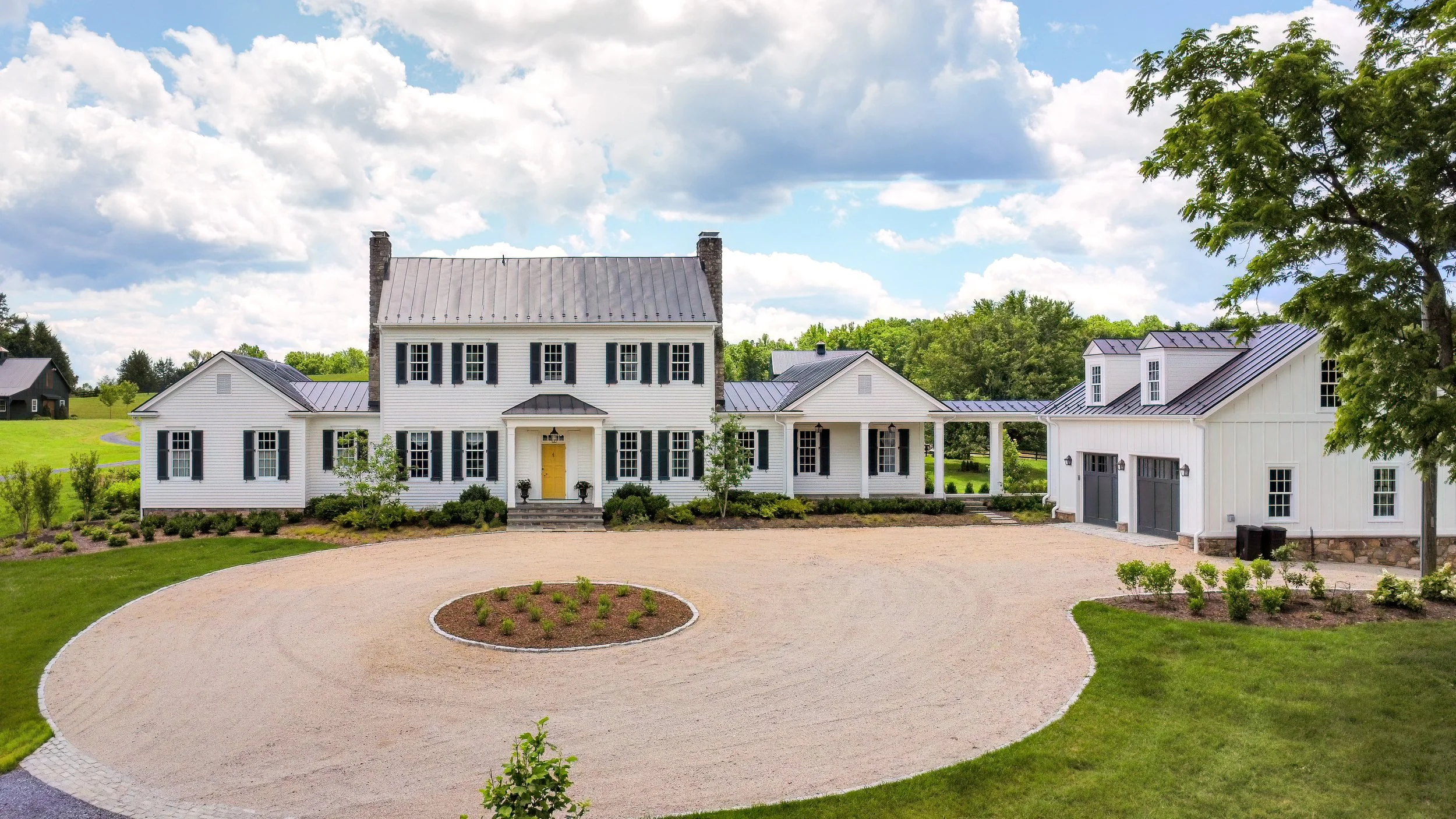 A large white two-story house with black shutters, a yellow front door, and a gray metal roof. Attached to it is a white garage with black doors, also with a gray metal roof. The house is surrounded by manicured lawns, bushes, and a circular gravel d