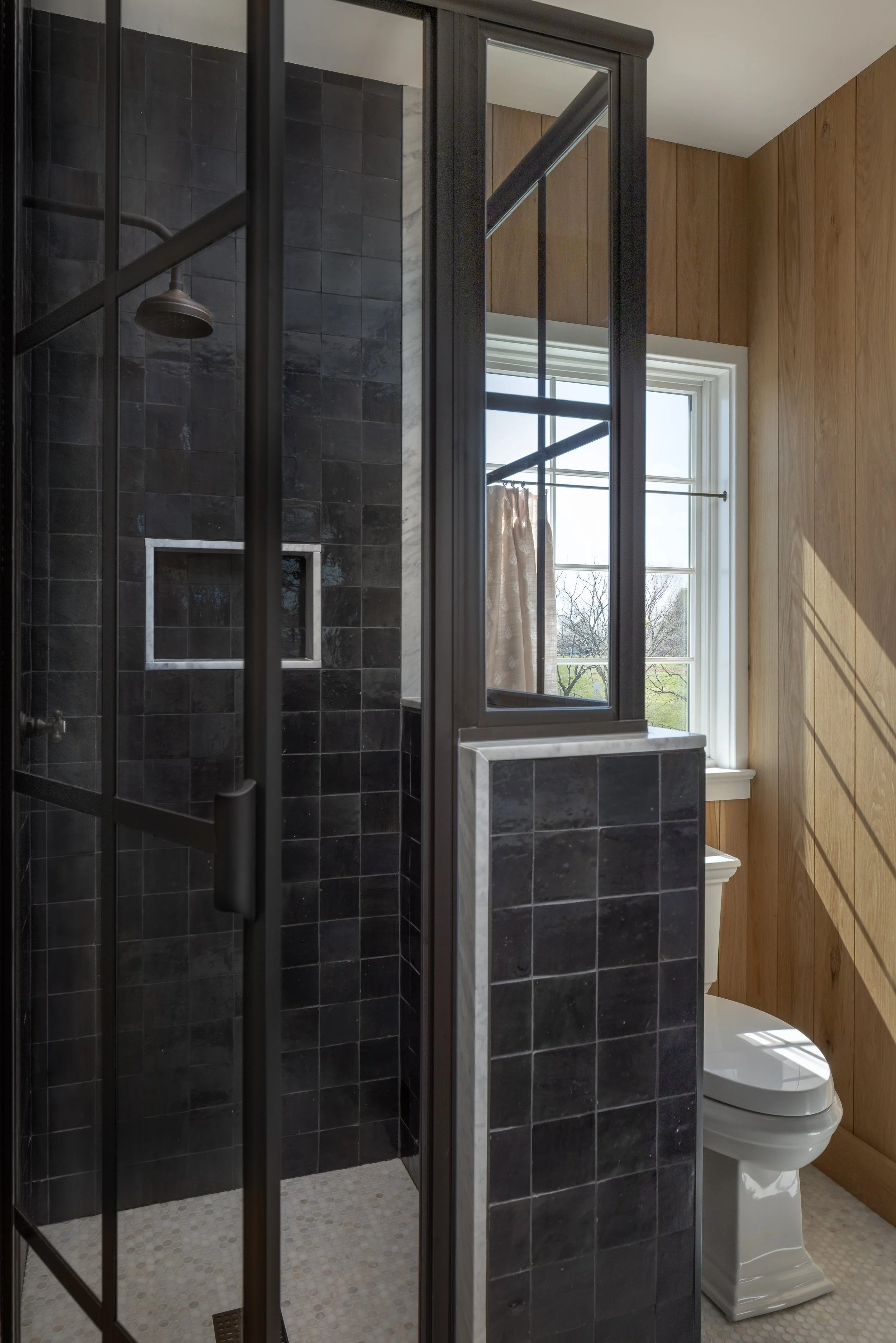 Small bathroom with a black tiled walk-in shower and a separate toilet area divided by a half wall with black framing. There is a window with natural light shining in, and wooden wall paneling on one side.