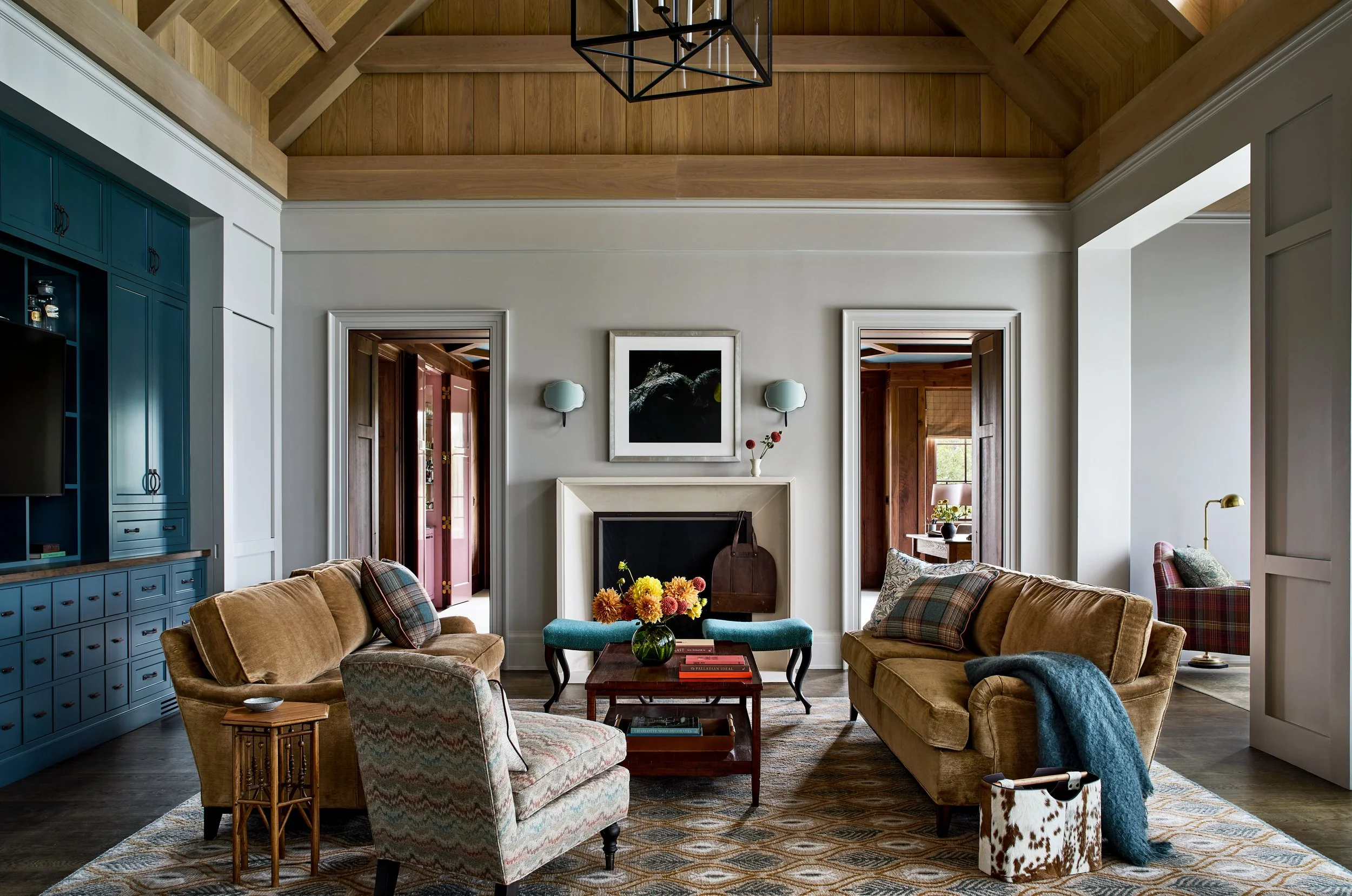 Living room with beige sofas, an armchair, wooden floors, and a fireplace with a black screen. There is a blue built-in unit on the left, wall-mounted lights, and a framed artwork above the fireplace. Decor includes flowers, pillows, and a throw blan