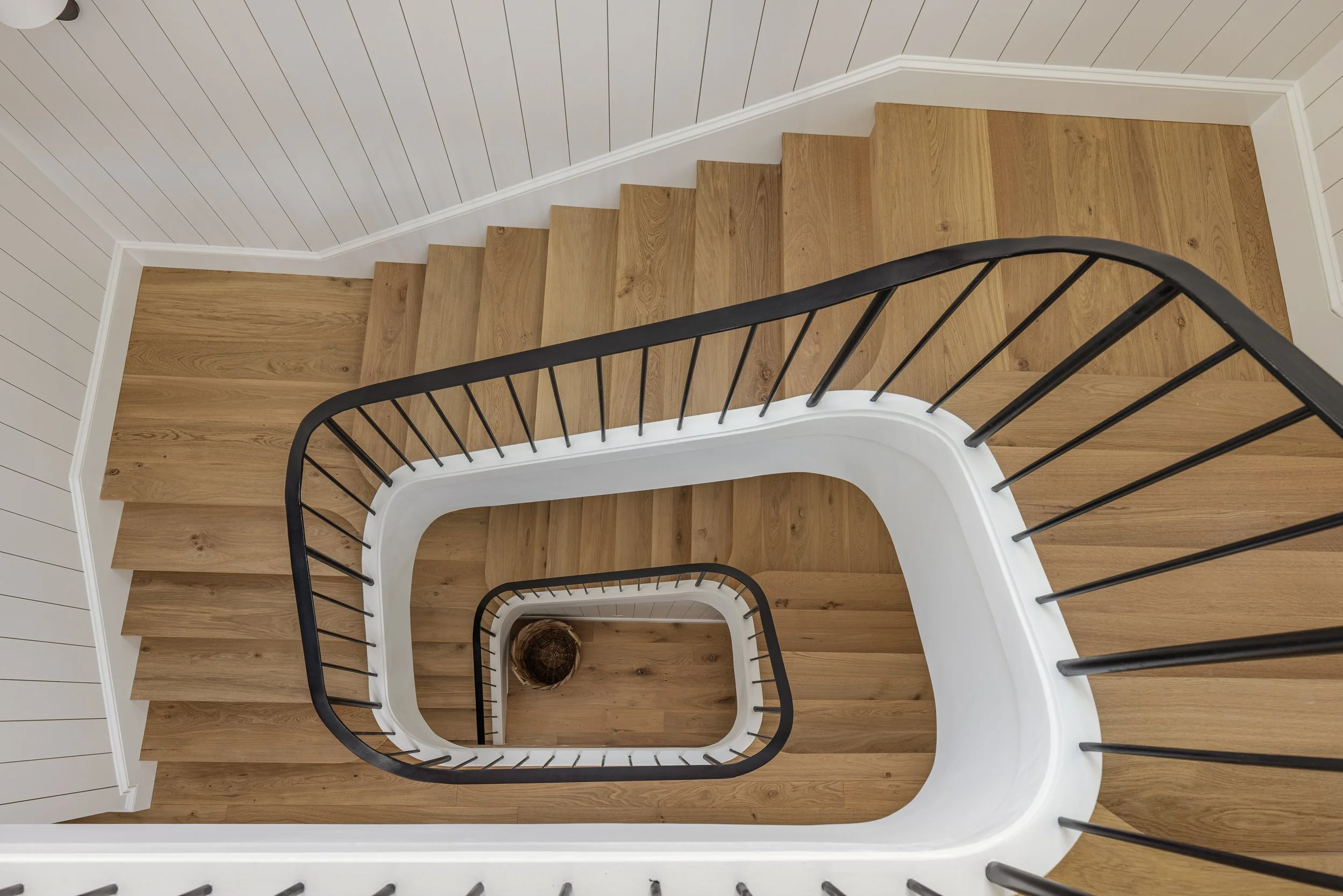 Overhead view of a spiral staircase with wooden steps, black handrails, white surrounding wall, and a small log at the bottom floor.