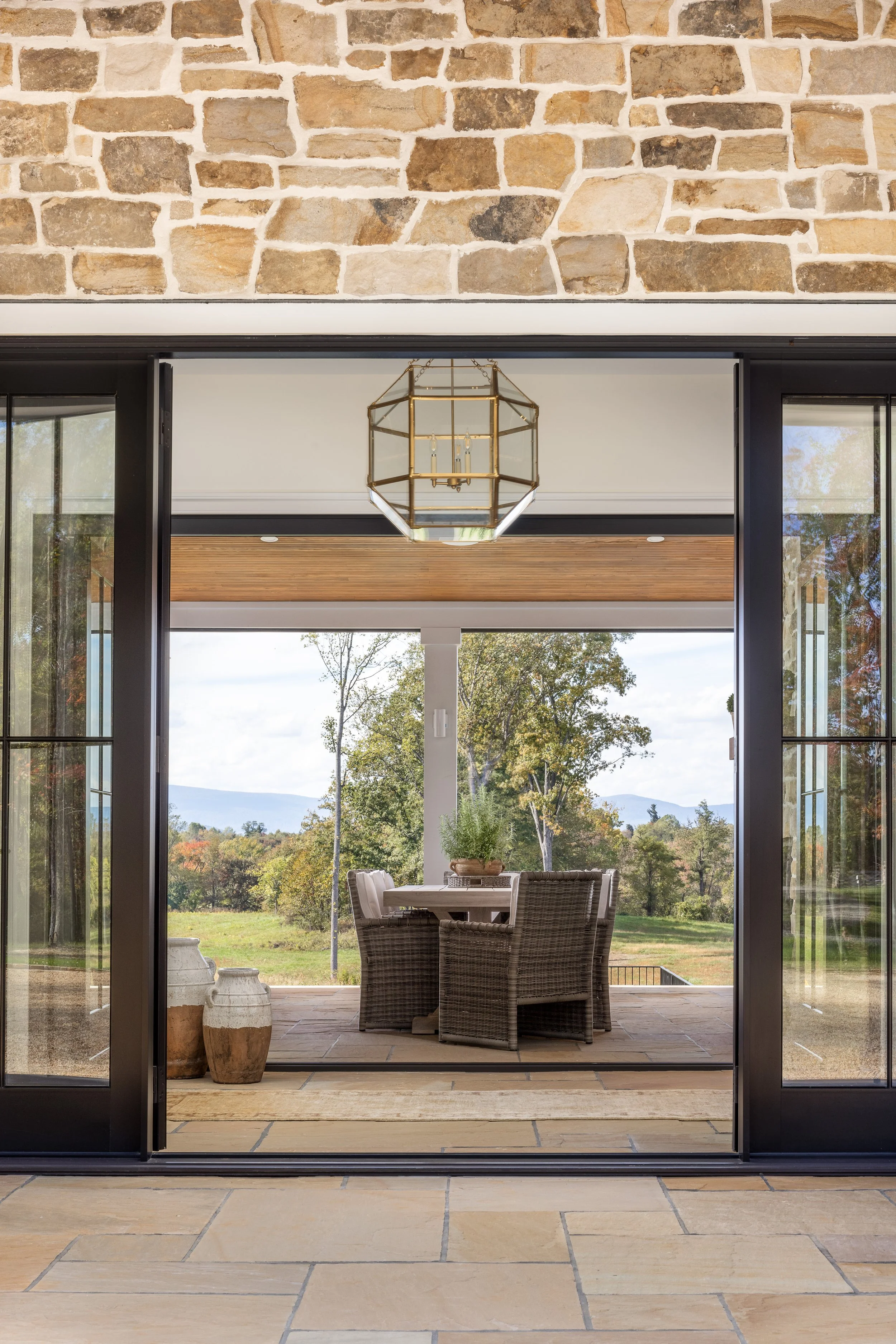 View of a patio with a round dining table and four wicker chairs, surrounded by lush green trees and mountains in the background, seen through open black-framed glass sliding doors, with a brass geometric chandelier hanging from the ceiling.