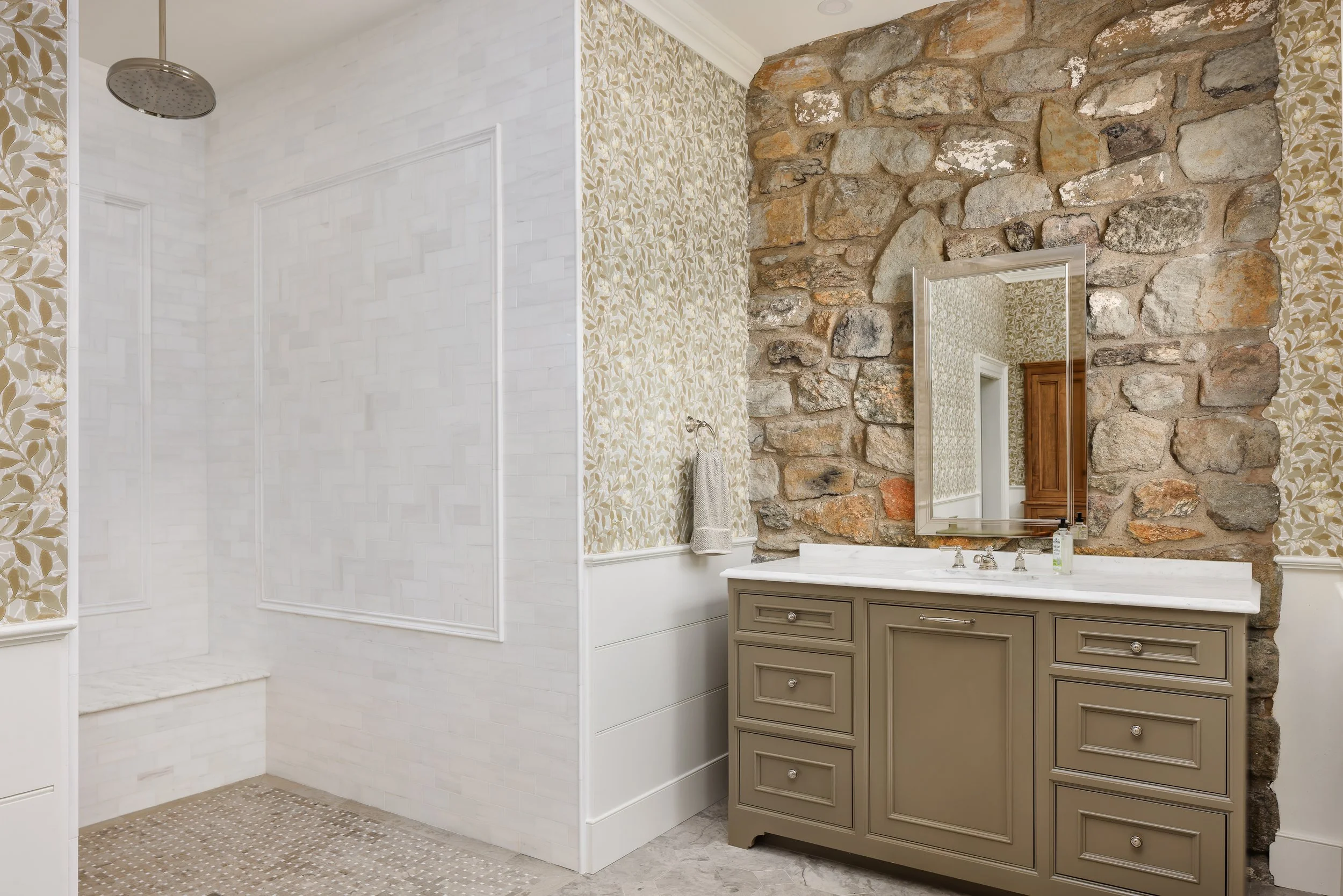 A bathroom with a walk-in shower, white tile walls, and a beige vanity with a mirror, with a stone wall and patterned wallpaper in the background.
