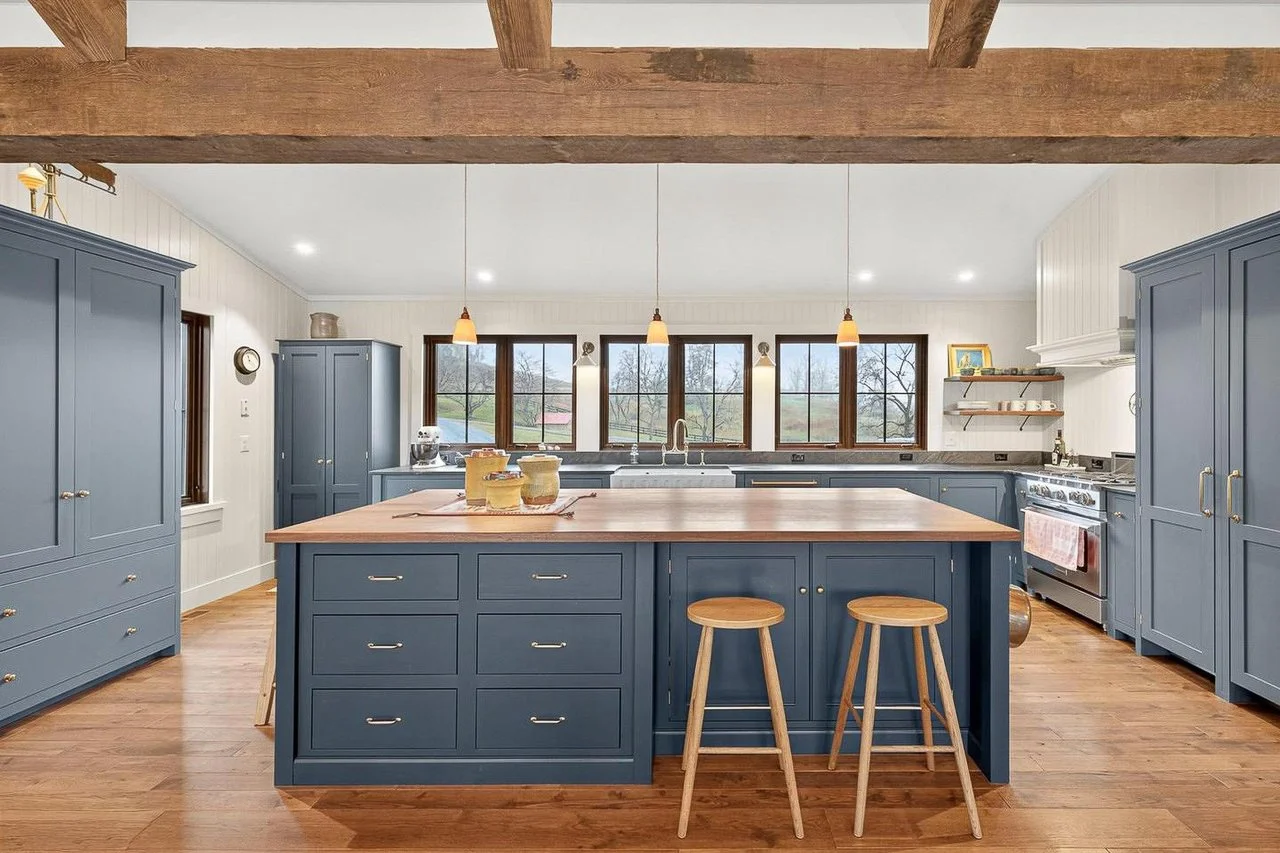 A spacious kitchen with blue cabinets, a large central island with a light wooden top, and a window view of trees and grass outside.
