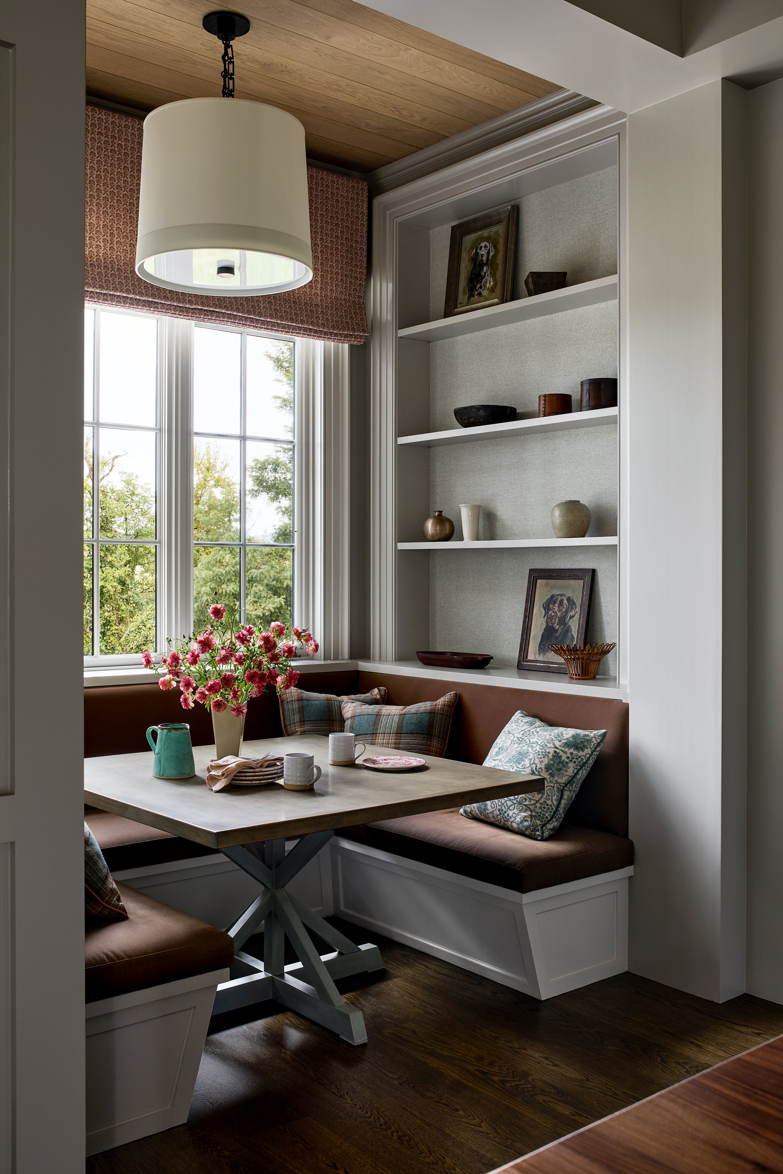 Cozy corner kitchen nook with window, built-in bench, and wooden table, decorated with flowers, mugs, and pillows.